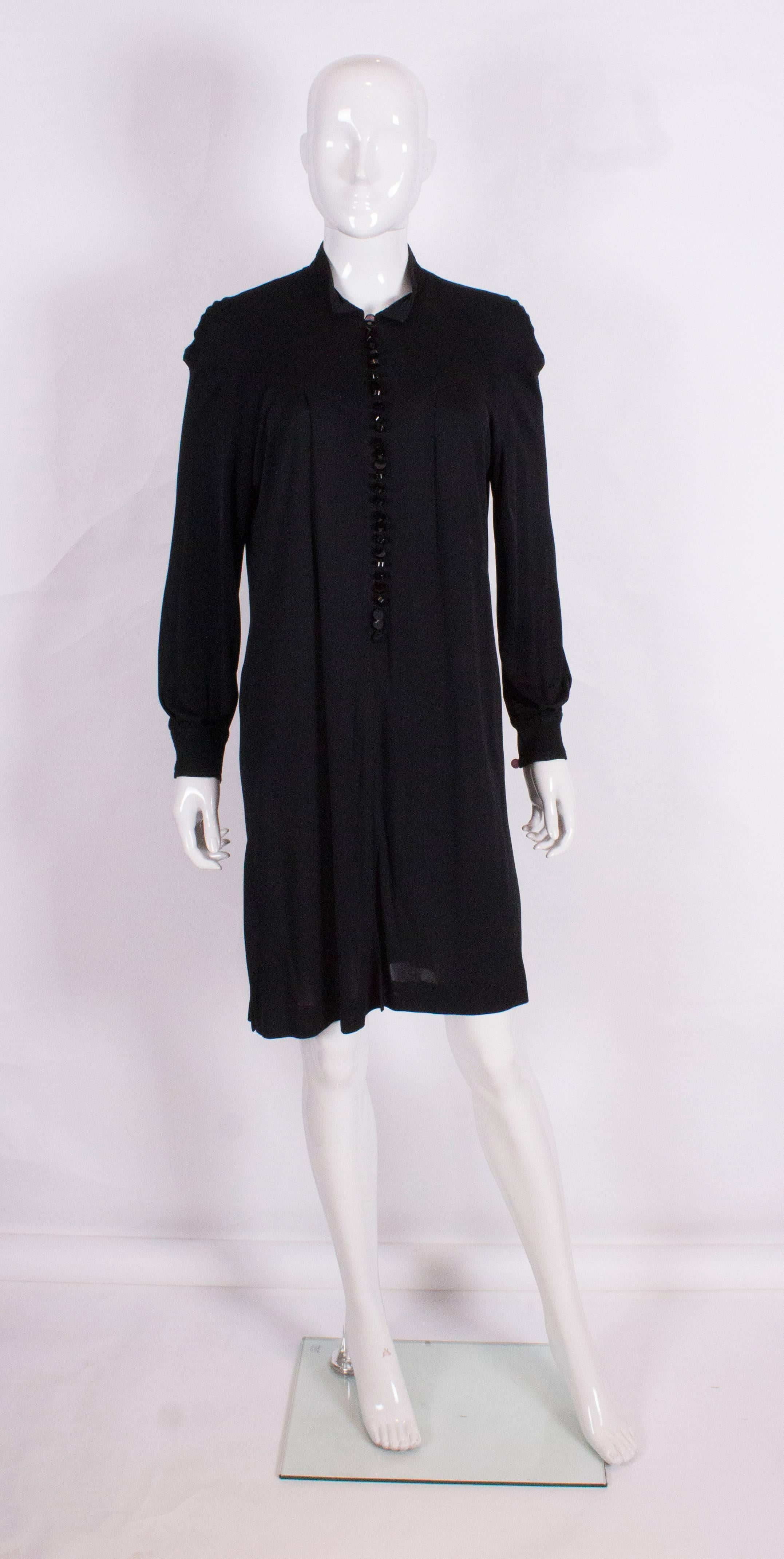 A chic tunic dress by Jean Muir.The black dress has a stand up collar with stitch detail,a button opening at the front,  single button on the cuffs, and pleats on the front and back bodice. It has a 9 1/2'' slit at each side and an 11 1/2 slit at