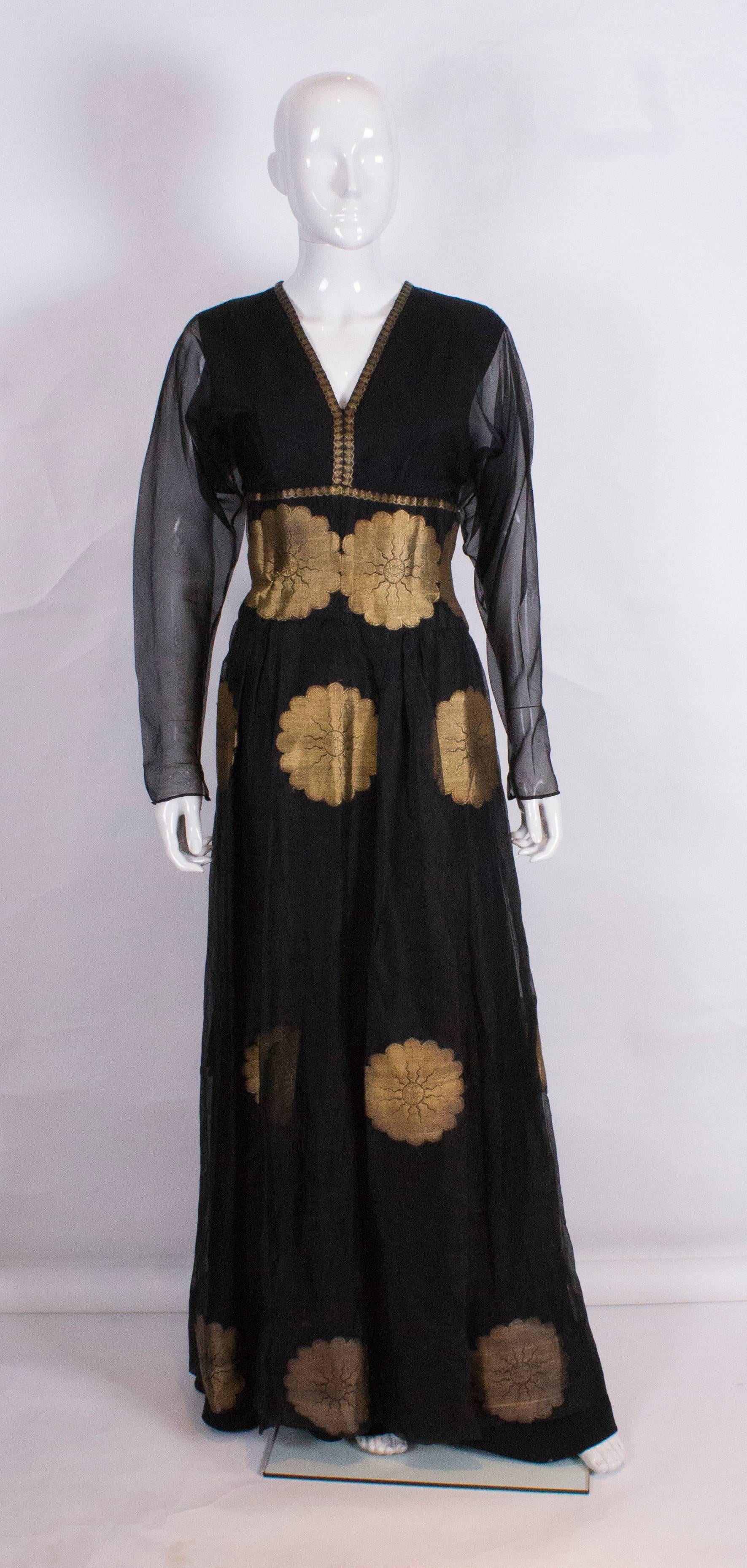 A chic evening gown in black silk organza over a black crepe underdress. The sleeves are sheer with popper and hook and eye openings.The dress has a v neckline , central back zip and wonderful gold lame flower detail.
