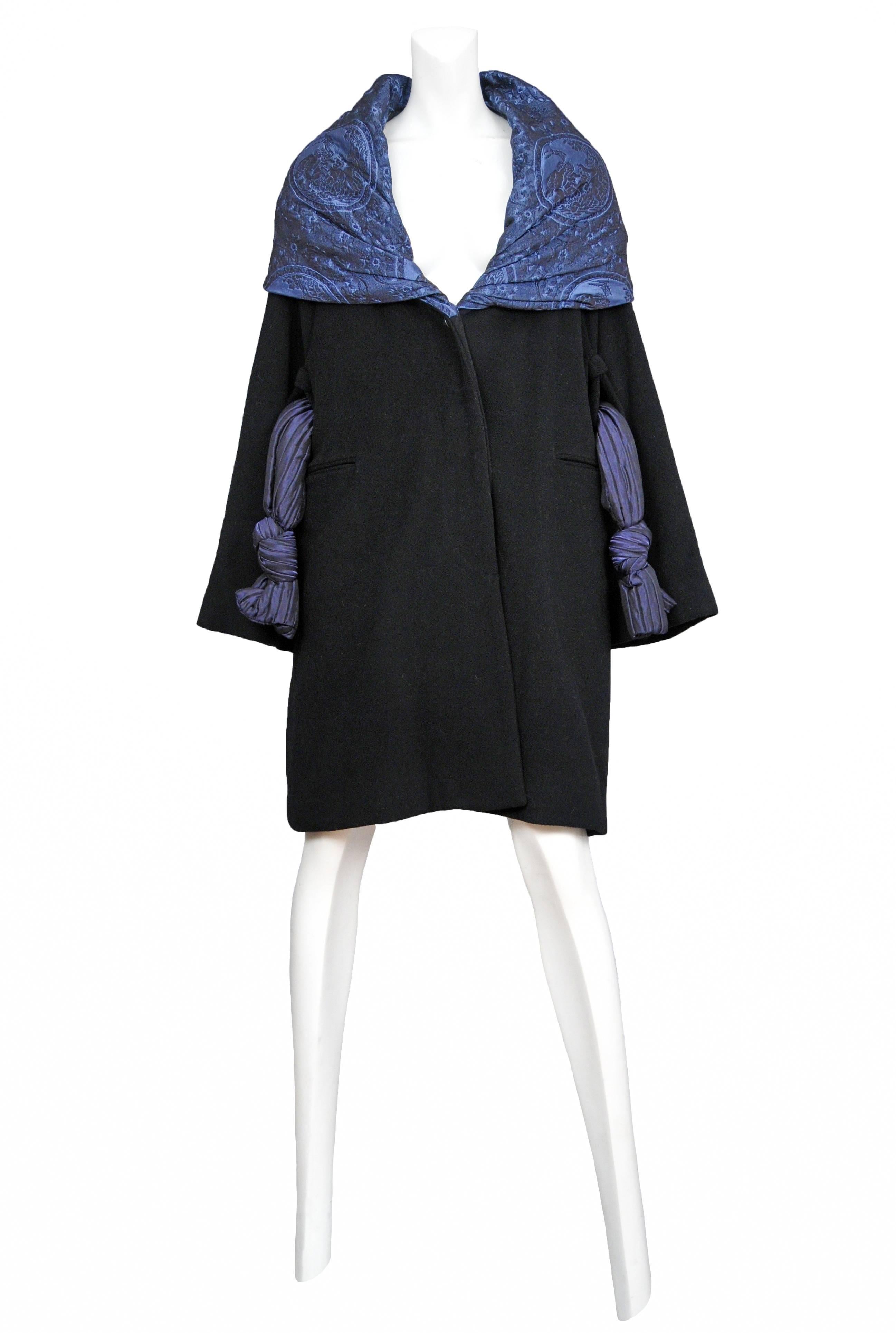 Vintage Romeo Gigli black wool knee length coat featuring a blue metallic embroidered shawl collar that fits over the shoulders, a metallic blue knotted belt that hangs freely on either side of the hips, and two side pockets.
Please inquire for