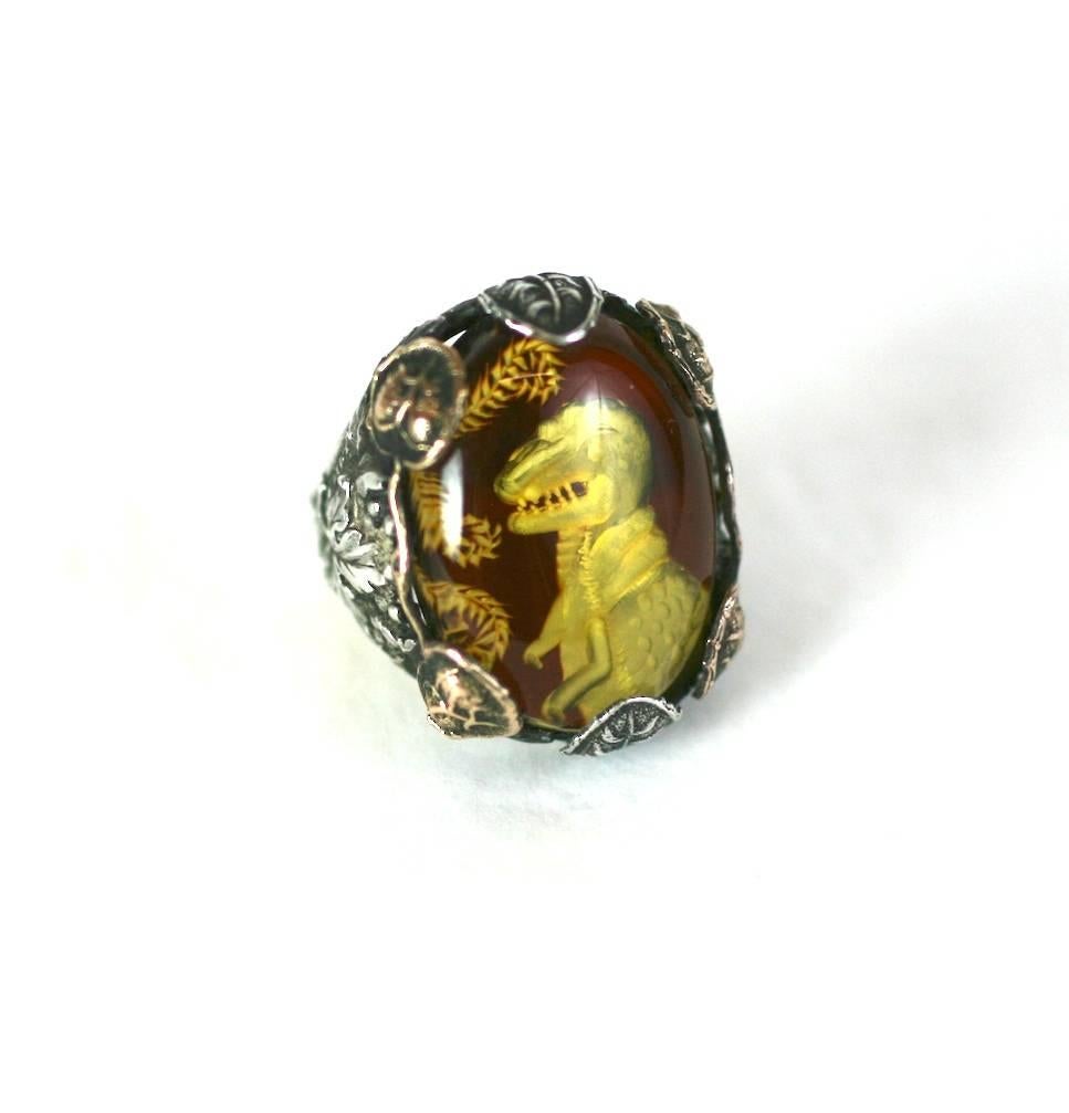 One of a kind ring with reverse carved amber cabochon of a dinosaur set into an antique Arts and Crafts setting, circa 1900. 
The sterling setting has elaborate leaf decoration on shoulders with both silver and coppery gold leaves used as
