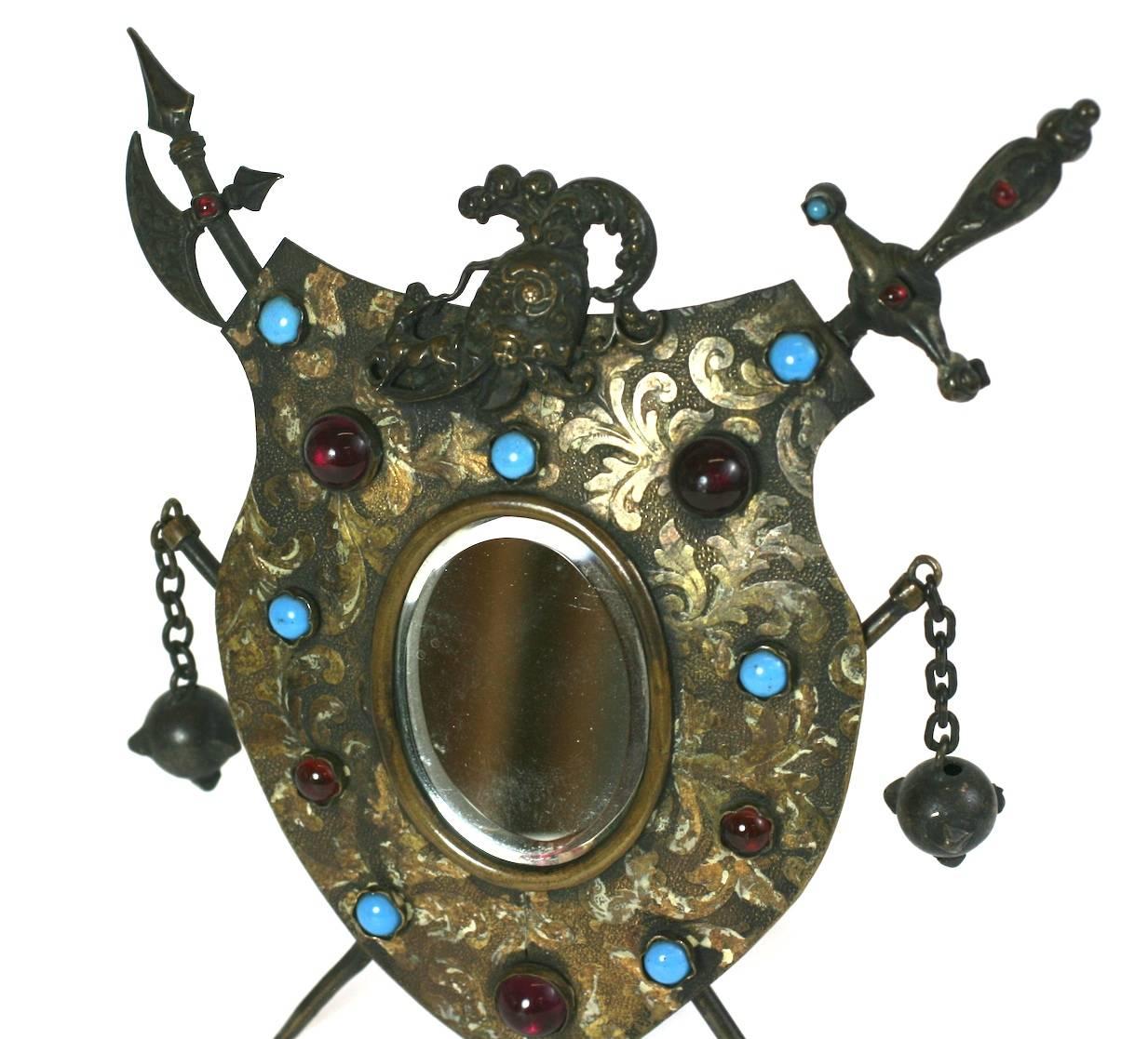 Super charming miniature mirror in a shield form with jeweled garnet and turquoise glass accents from the late 19th Century. Sword and Axe motifs form the cross legs with ominous spiked ball weaponry dangling from each side.
A knight's helmet caps