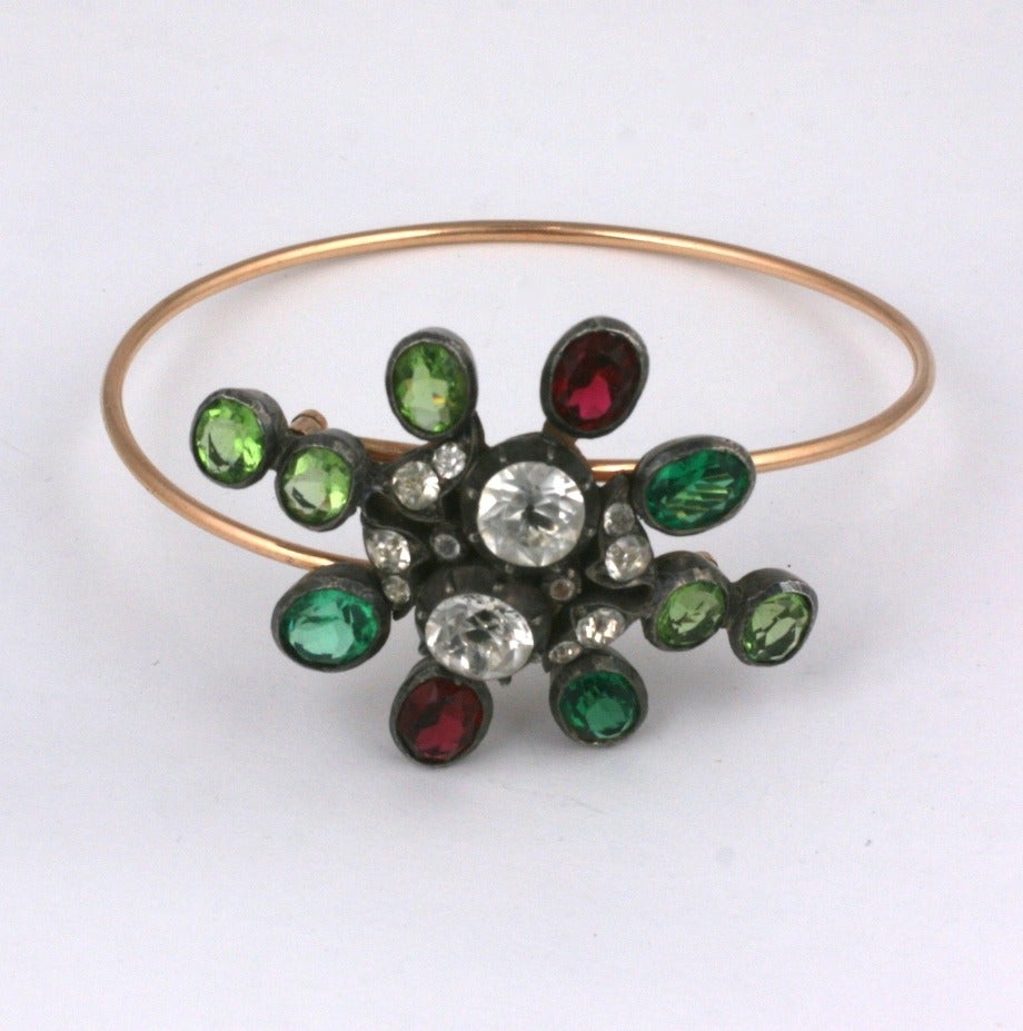 Unusual Early Paste Bracelet with a unique mechanism for entry. The central motif is set with red green and clear pastes on silver topped 14k gold. The wire bangle is set in gold filled metal. 
To open, the motif "slides" open sideways to