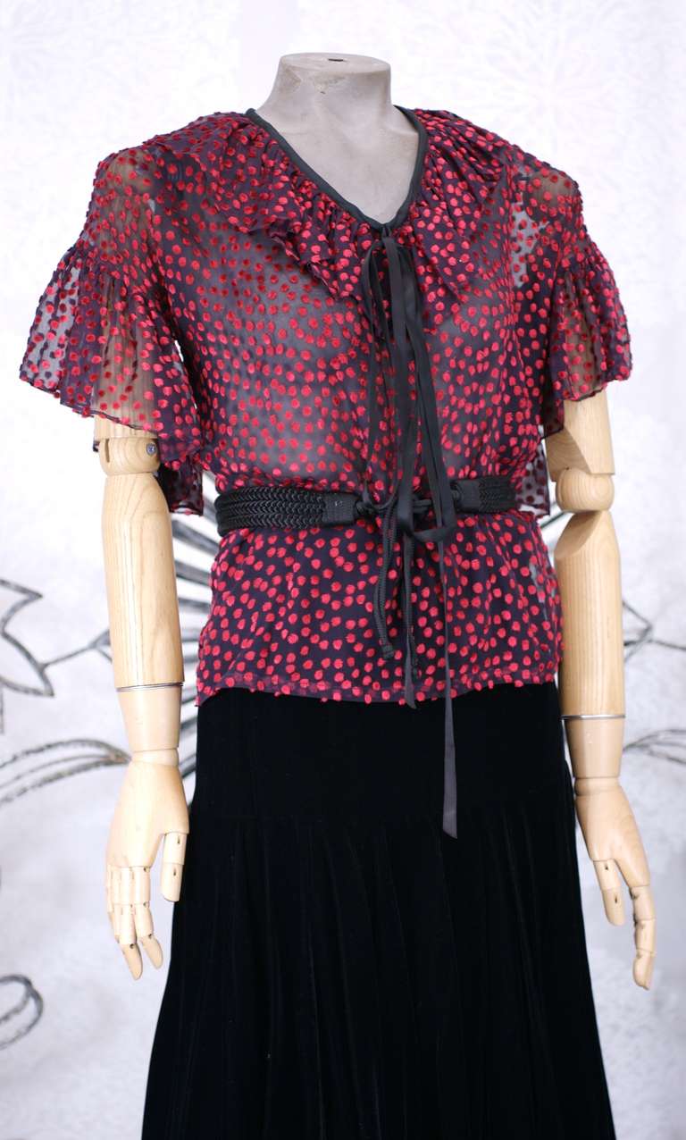 Yves Saint Laurent Silk Chiffon and Velvet Ensemble. Sheer black silk chiffon top with red coupe de velour polka dots rendered with ruffle trimmed sleeves and collar and a long black satin tie. 
Black cotton velvet skirt is simply cut with a hip
