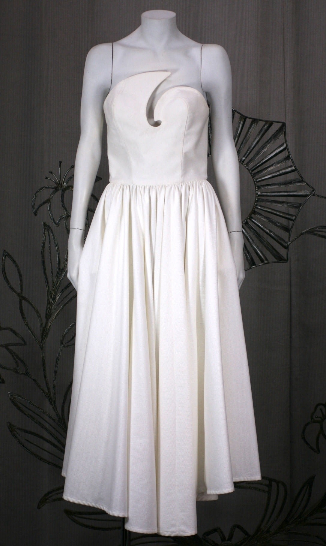 Thierry Mugler's dramatic white cotton pique sun dress with assymetrical swirl cut outs on strapless bodice with full dipping hem circle skirt. Bodice is boned for support and snaps up the back. Hem dips in front and has self belt with antique