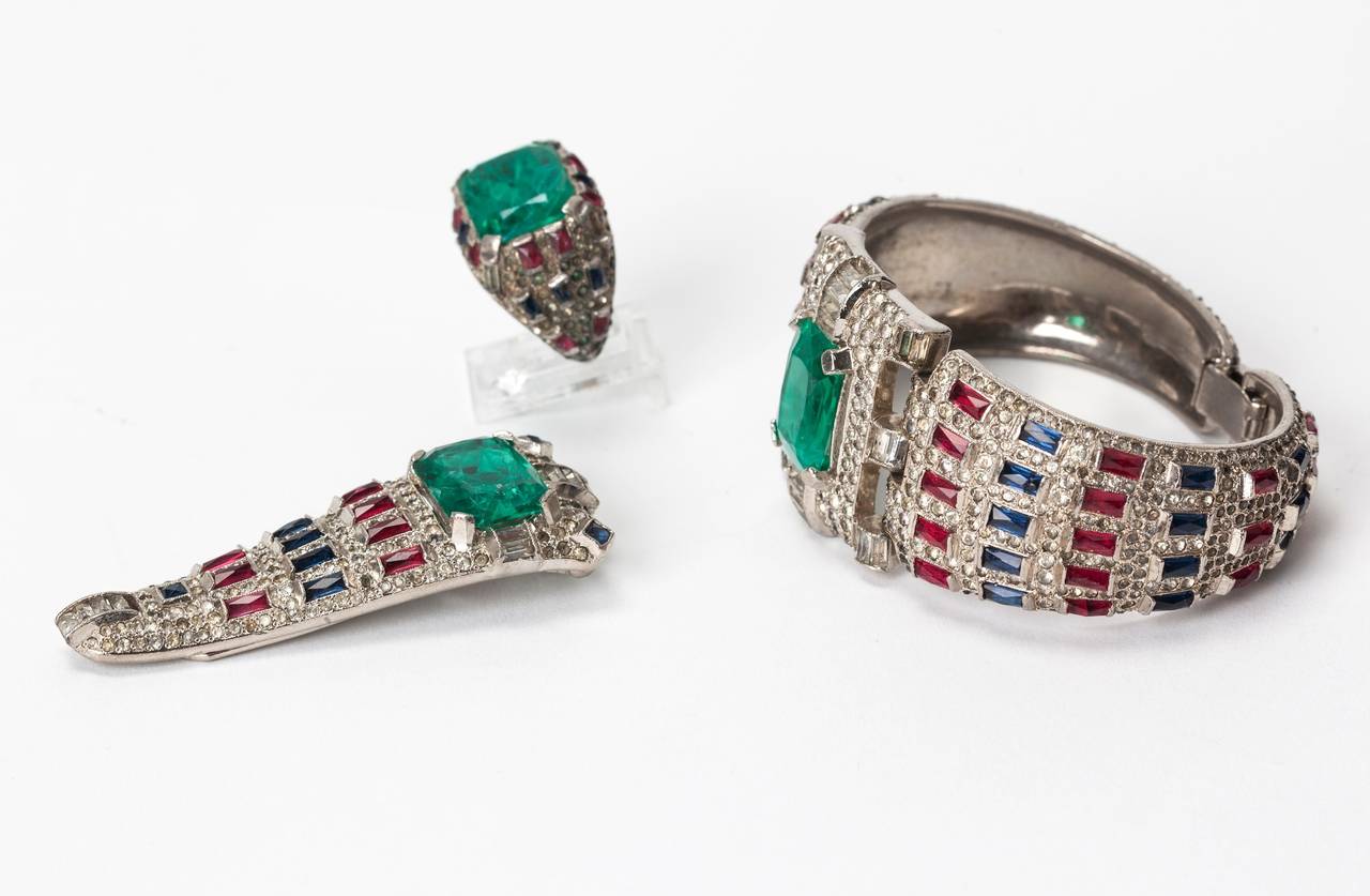 Important Early Art Deco Trifari Suite composed of cuff, dress clip and cocktail ring. Each design centers around a large faux emerald and is studded with pave work and faux ruby and sapphire baguettes. 
Exquisite high quality manufacture and design