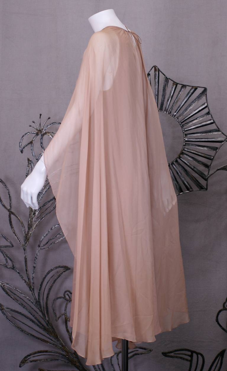 Halston Silk Chiffon Cape Dress in nude silk chiffon. 3 ply silk chiffon bias cut sleeveless sheath with an attached cut away cape which descends to full length in the back. Silk self tie at neck knots at back for neck for closure. 
Cape is split in