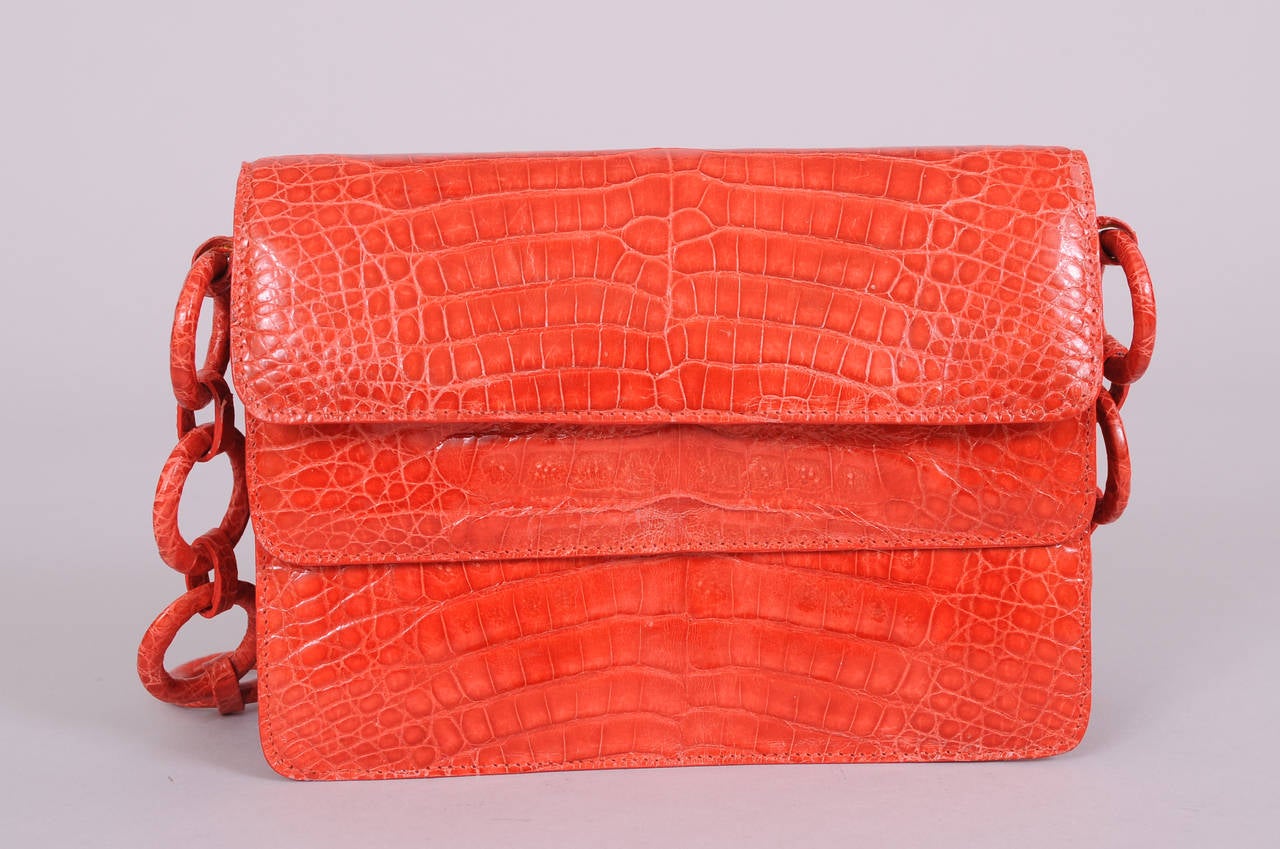 A colorful coral colored crocodile skin bag is accented with a strap made from crocodile covered rings. There are two sections, each with a hidden magnetic closure, and pale beige suede lining. The bag is in excellent condition and appears