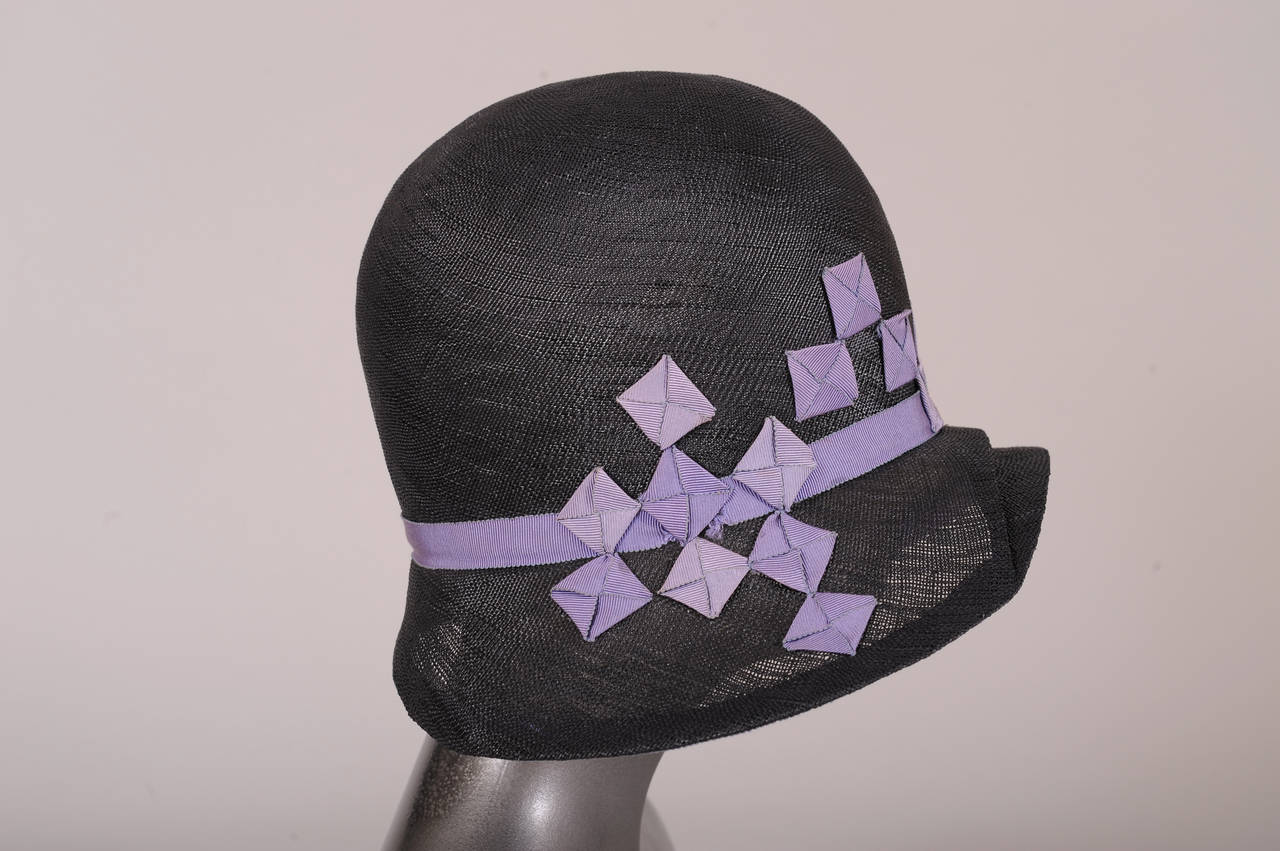 A classic black straw cloche hat originally designed by famed Paris milliner Agnes, an authorized copy, is trimmed with lavender grosgrain ribbon. The hat has a ribbon hat band and clusters of folded ribbon squares sewn to the crown and brim. The