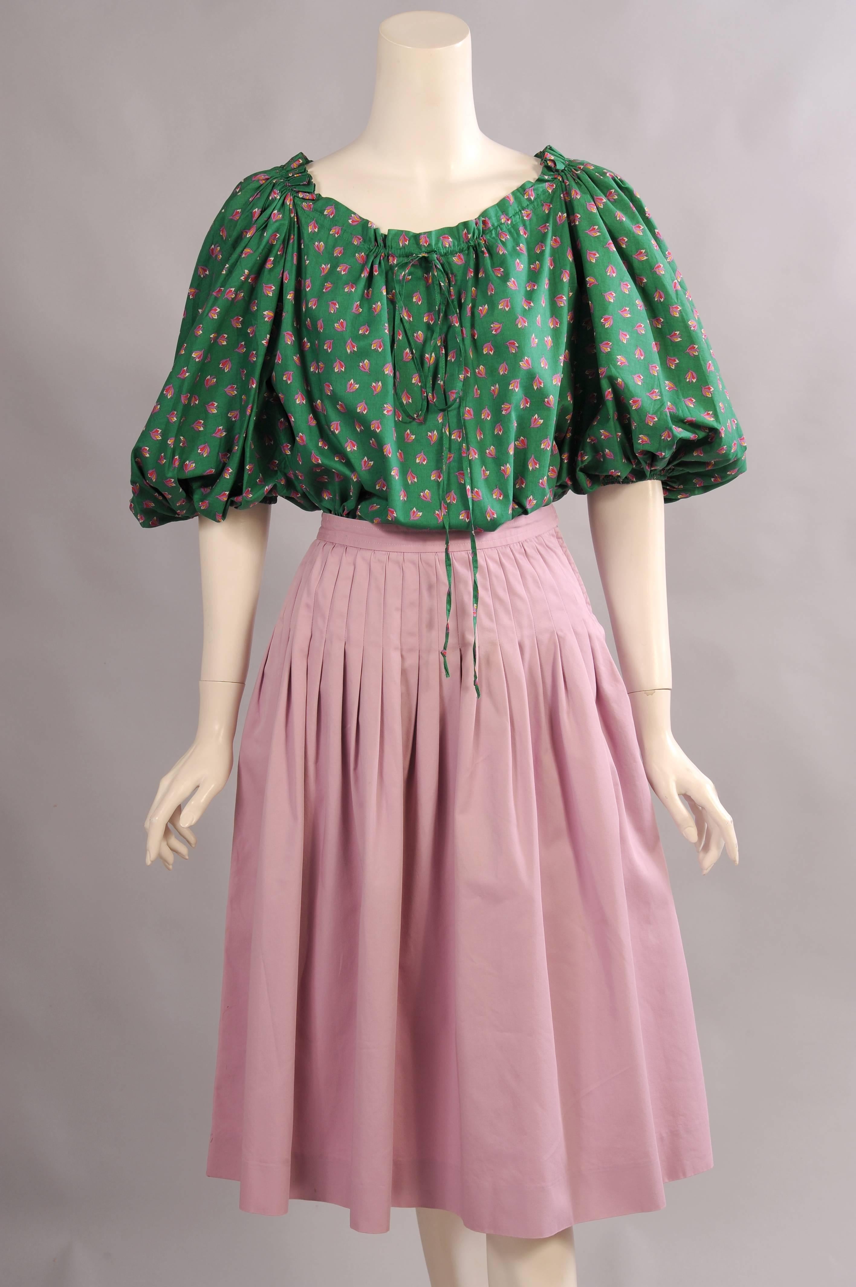 A bright green peasant style cotton blouse with a lavender floral design has a gathered drawstring neckline and full sleeves with elastic. The coordinating lavender cotton skirt has a natural waistline, stitched down pleats and two pockets concealed