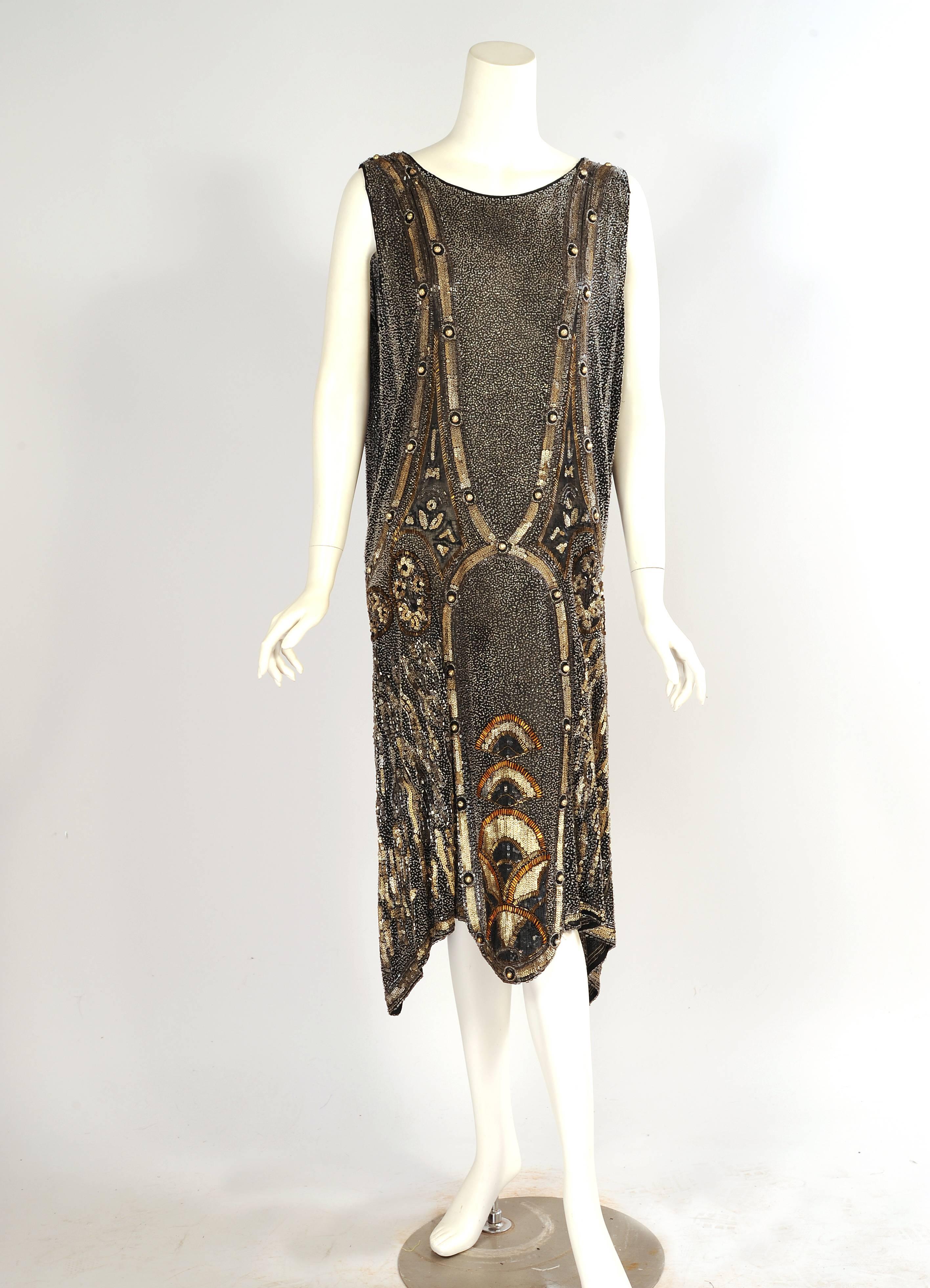 This stunning Art Deco flapper dress is is completely beaded. The body of the black cotton dress is covered with tiny clear beads. Silver bugle beads outline the graphic design which is made with textured matte gold sequins and gold blister pearls. 