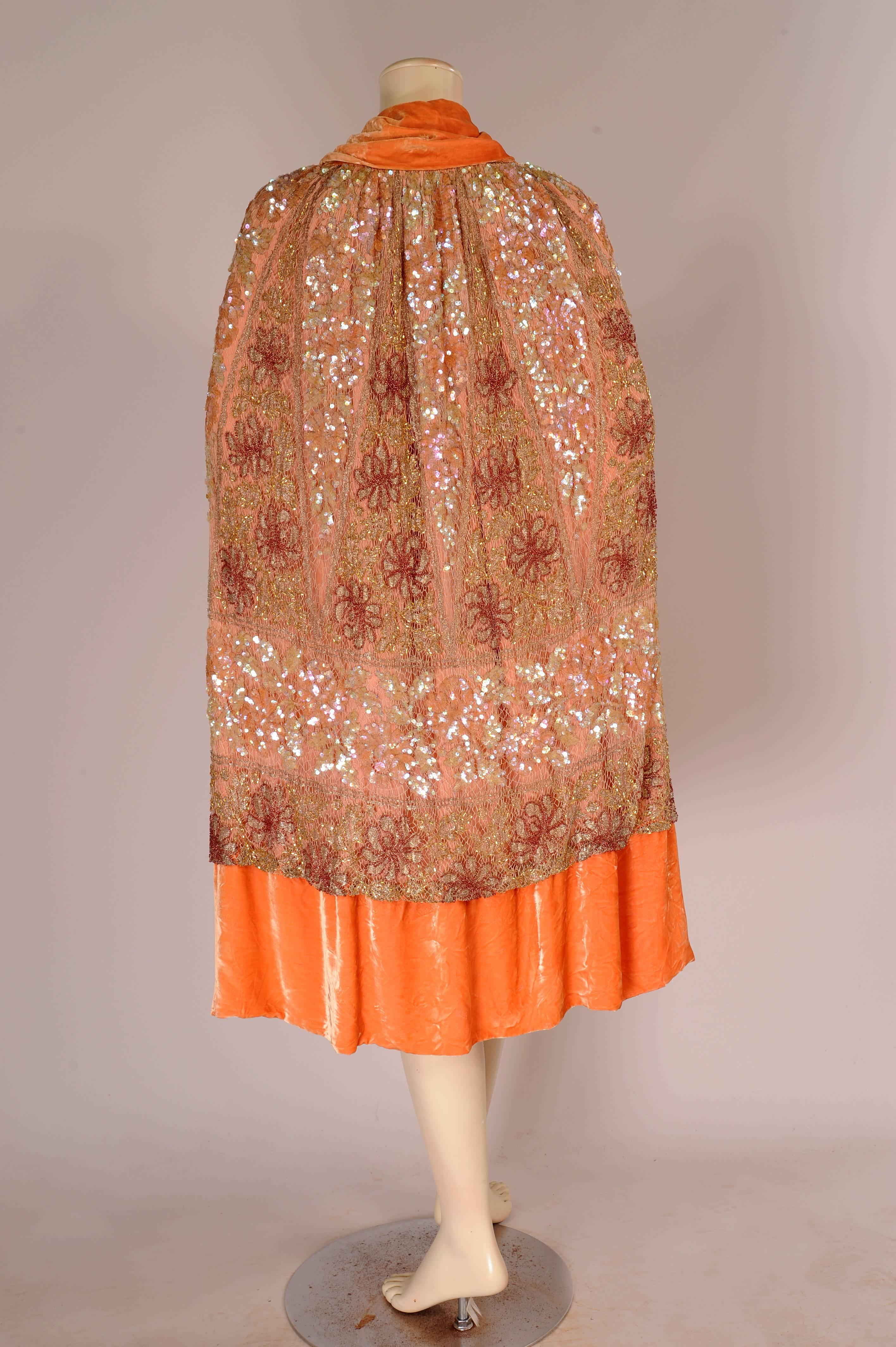 A striking example of 1920's clothing, this evening cape is made from apricot colored silk velvet. It has a large decorative panel of beadwork and metallic lace covering almost all of the velvet except for the border and scarf tie at the neck. The
