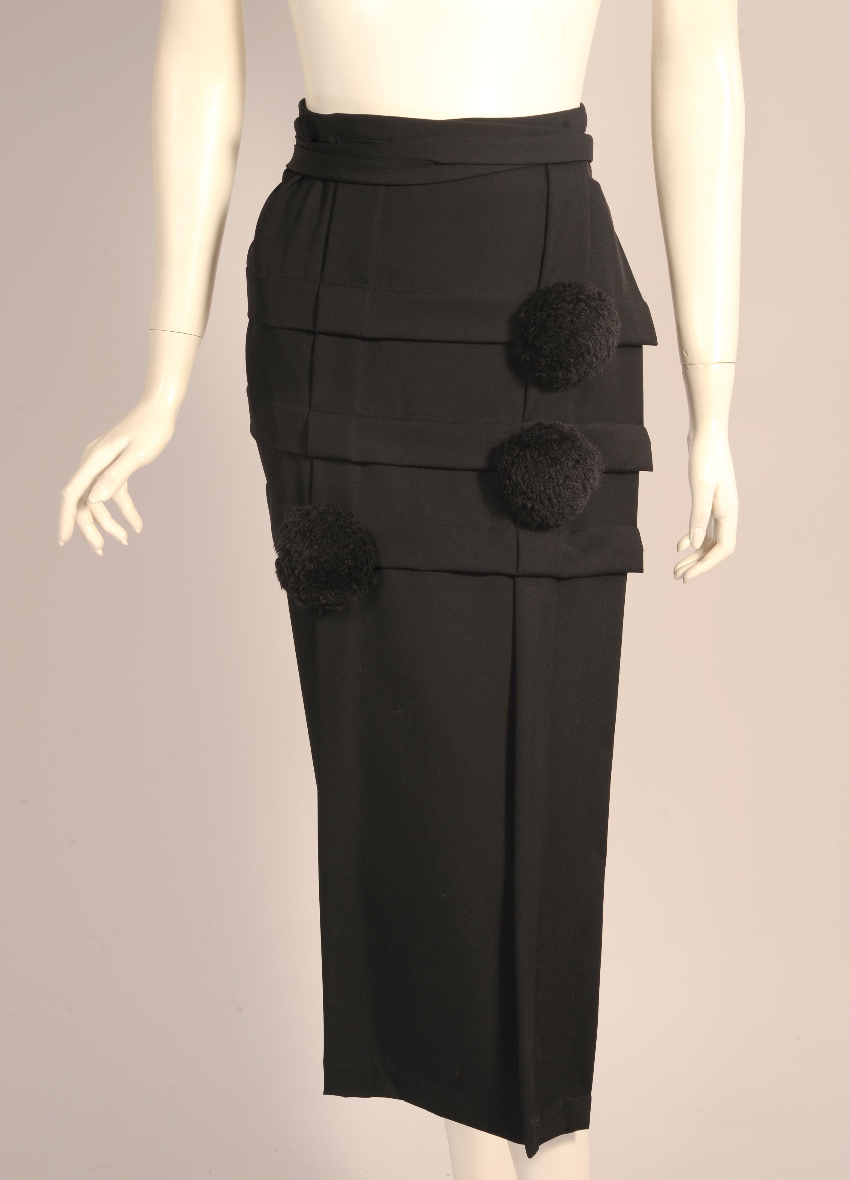 This sleek black wool rectangular wrap skirt has a gri9d of wide pleats at the front. Two are vertical from waist to hem and three are horizontal at the top of the skirt. They are embellished at their intersections with three large black wool