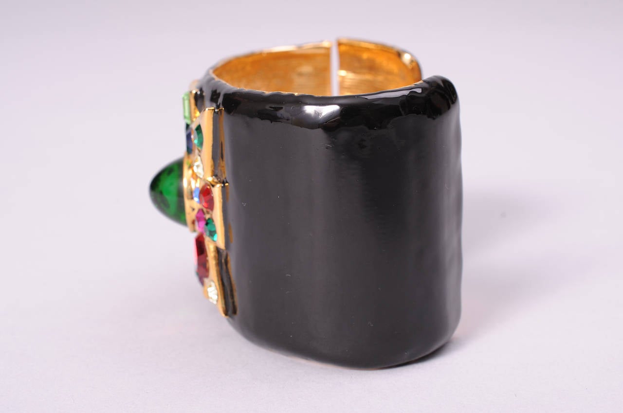 This stunning black enamel cuff has a striking Maltese cross set with faceted and cabochon stones. A large green cabochon  sits in the center surrounded by an array of smaller stones. The bracelet has a hinge on one side so it is very easy to slide