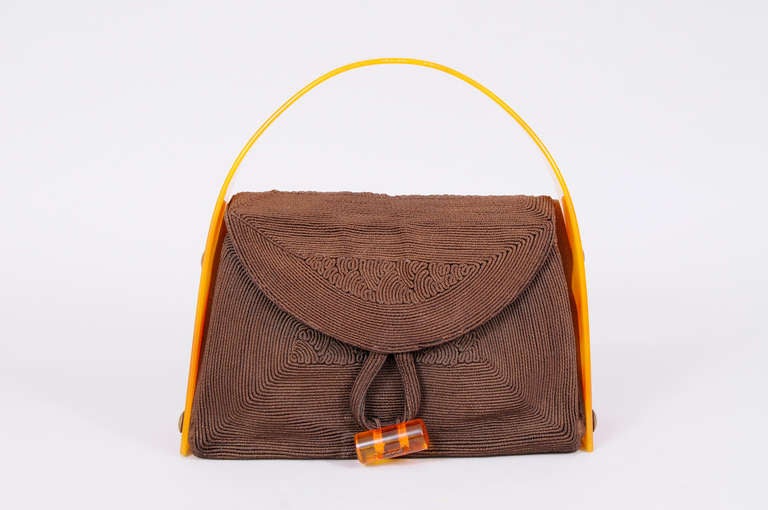 A fabulous Bakelite handle arches over this brown Corde handbag. It is attached to the sides with brass buttons. There is a Bakelite cylinder and loop closure, and there are two pockets inside. The bag is in excellent