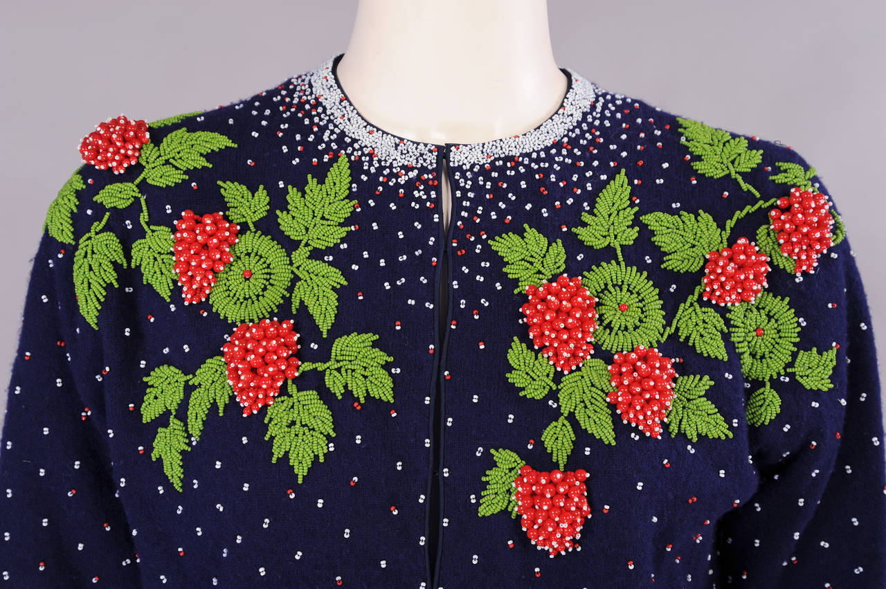 Luscious three dimensional red strawberries on a beaded green vine turn this navy blue cashmere cardigan into something quite special. The berry motif is repeated on the cuffs as well. The sweater is edged with red and white beads which are also