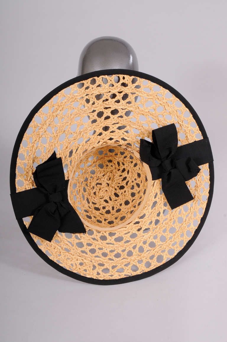 A large brimmed hat in open weave natural straw is trimmed with black grosgrain ribbon. The ribbon edges the brim and wraps over the brim in two places. On the under side of the brim there are two large signature Chanel bows. It is in excellent