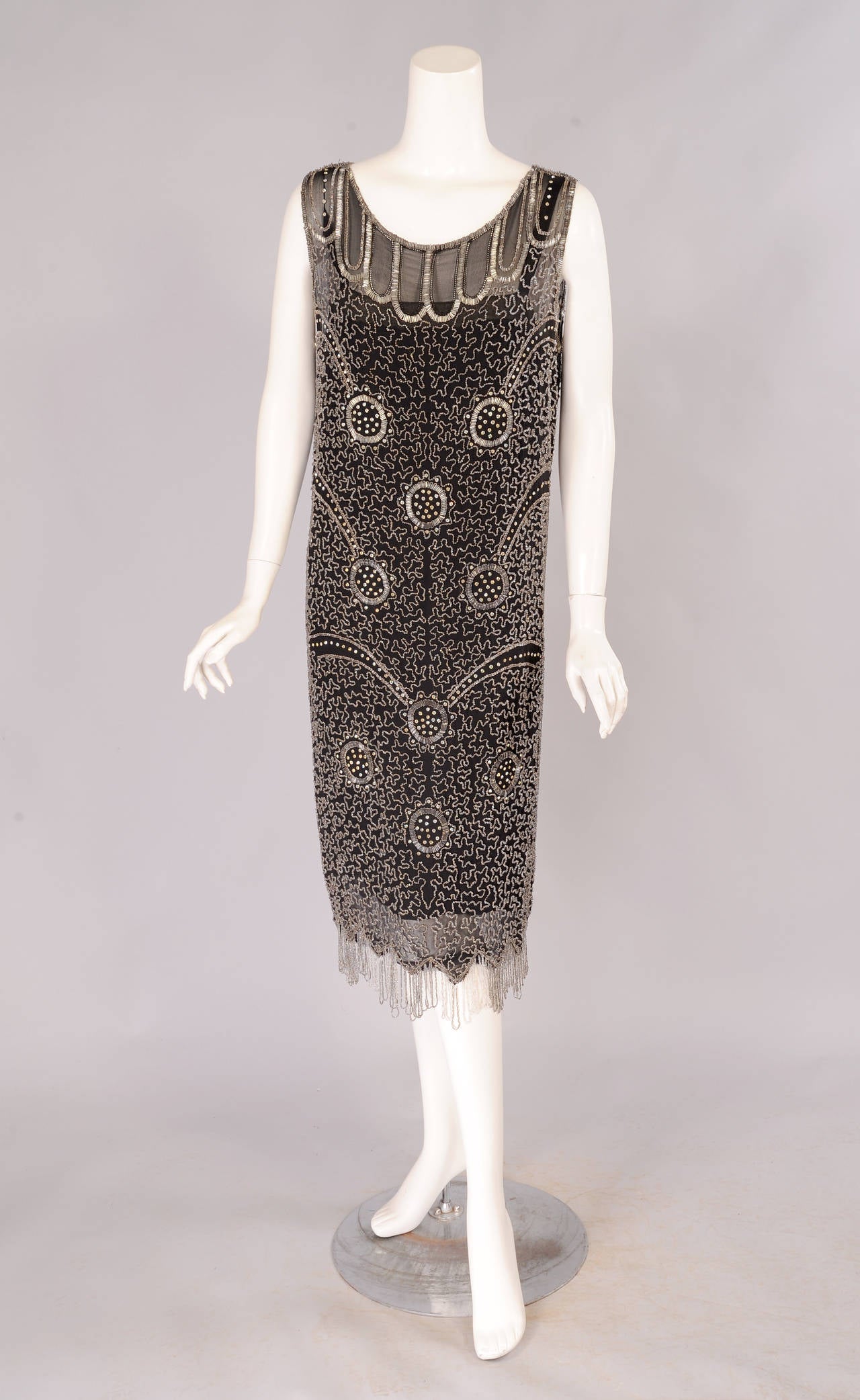 Black silk is hand beaded with a bugle beaded design at the neckline and a vermicelli pattern in caviar beads on the body of the dress. This is further embellished with prong set rhinestones in circular motifs. The dress has a looped beaded fringe
