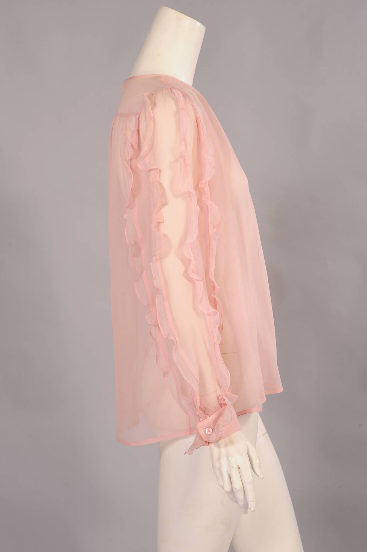 Pink silk georgette is embellished with rows of ruffles running the length of the sleeves. The French cuffs are also edged with ruffles. The blouse has a single button at the back above a deep opening. The blouse is in excellent