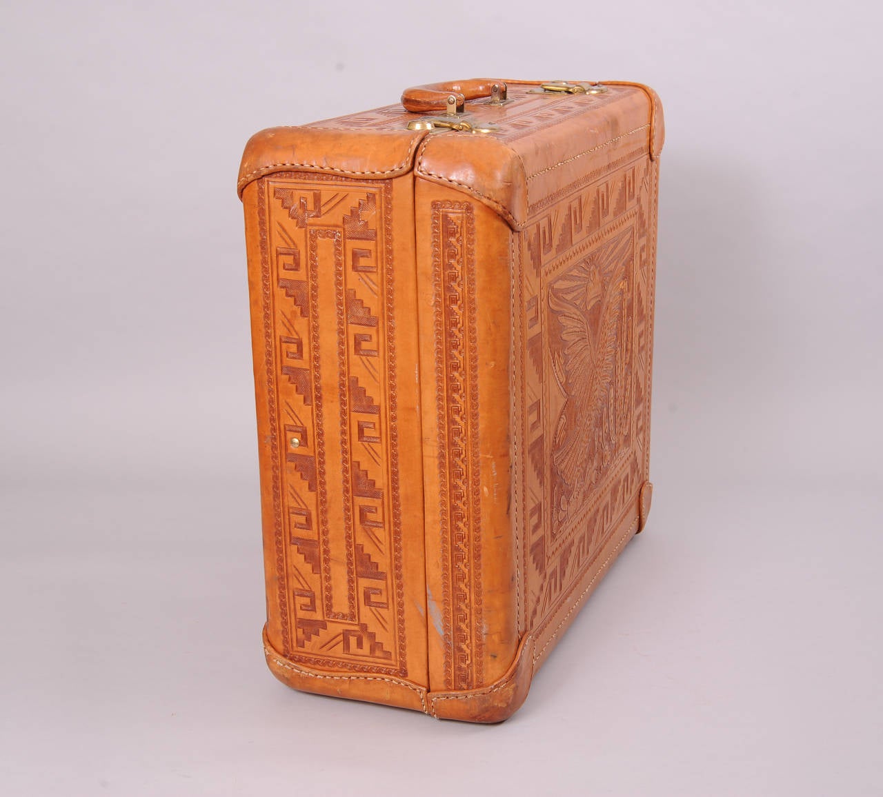Covered with exquisite hand work, this leather suitcase, circa 1950, has a large bird in a cactus garden on the front. This panel is surrounded by a border of scallops, bold geometric designs, and decorative waves. This border is repeated on all