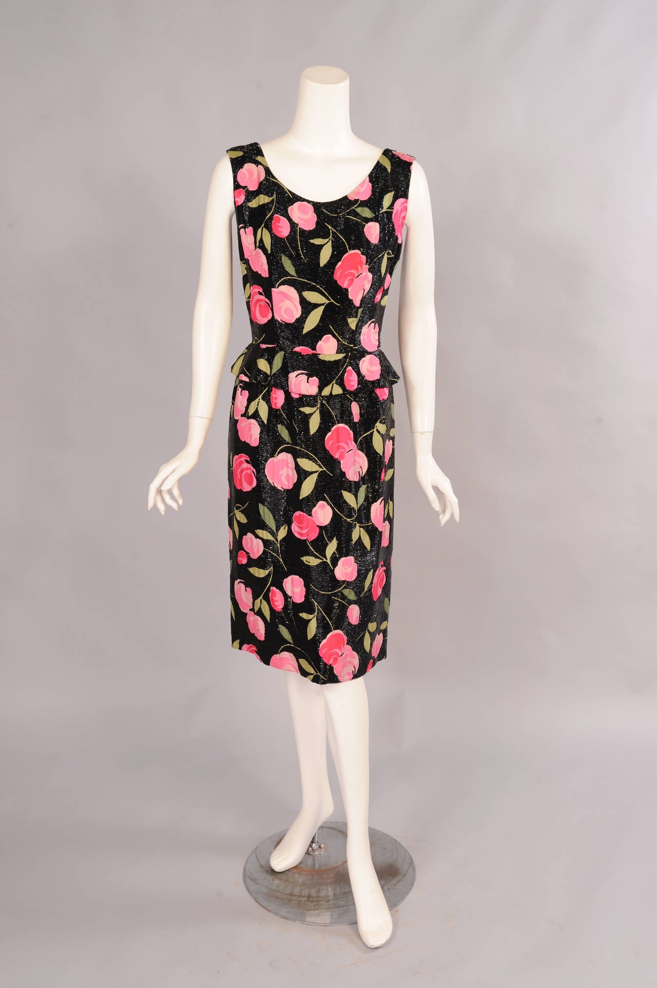A charming pink and green floral print on a black silk background is covered in tiny black bugle beads. The dress has a peplum at the waist and a center back zipper. It is fully lined with a combination of pink organza and black taffeta. It is in