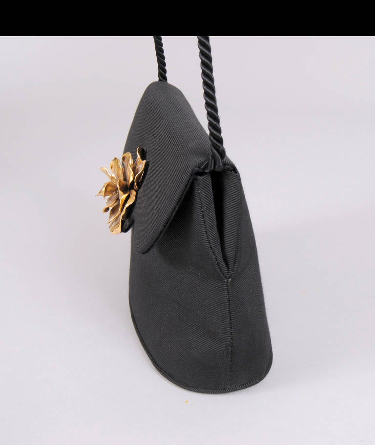 In pristine condition, this bag appears unused. The silk faille bag is accented with a large three dimensional gold toned flower at the closure. A black silk cord can be used as a shoulder strap or tucked inside. The bag is lined in black satin and