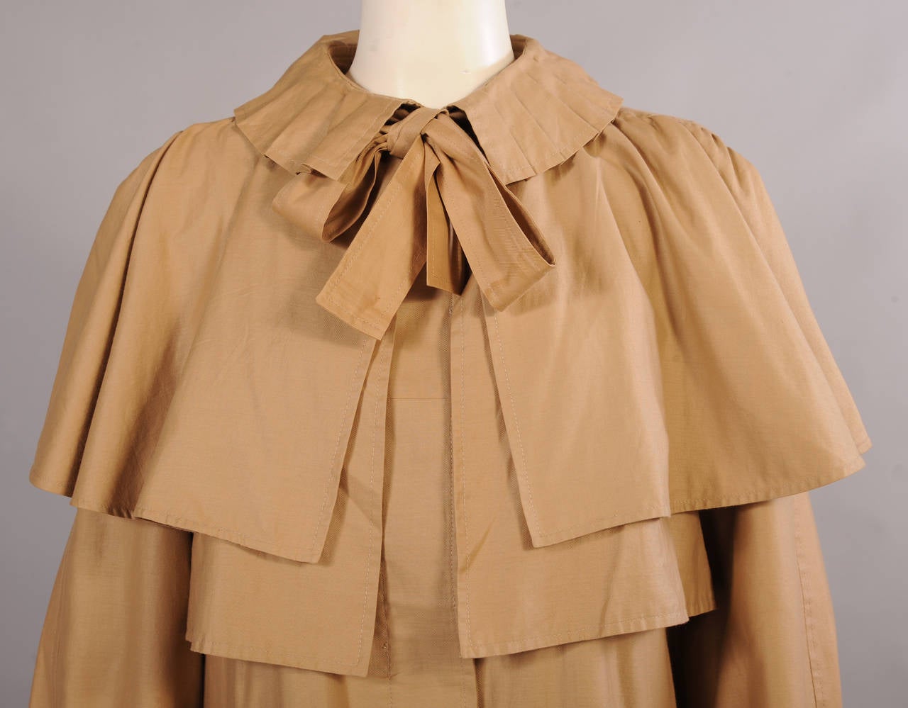 A ruffled collar and an attached capelet add interest to this cotton coat from the 1970's. The bow at the neckline is really just the matching belt tied under the collar. The coat has hidden buttons, D rings on the cuffs and two pockets. It is in