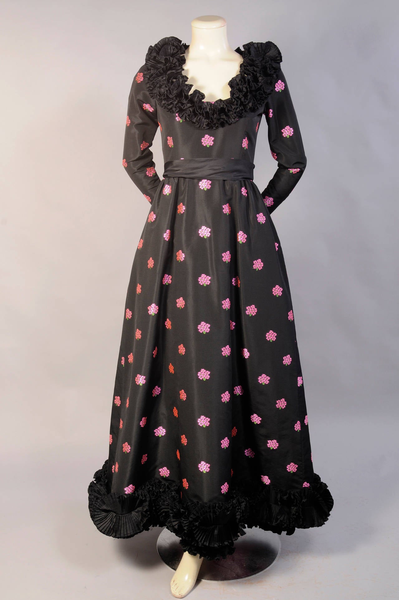 This elegant evening gown designed by YSL in the early 1970's is a striking combination of woven pink and green floral bouquets, black silk and a fabulous shocking pink ruffled petticoat. The dress has a low ruffle edged neckline and a deep ruffle