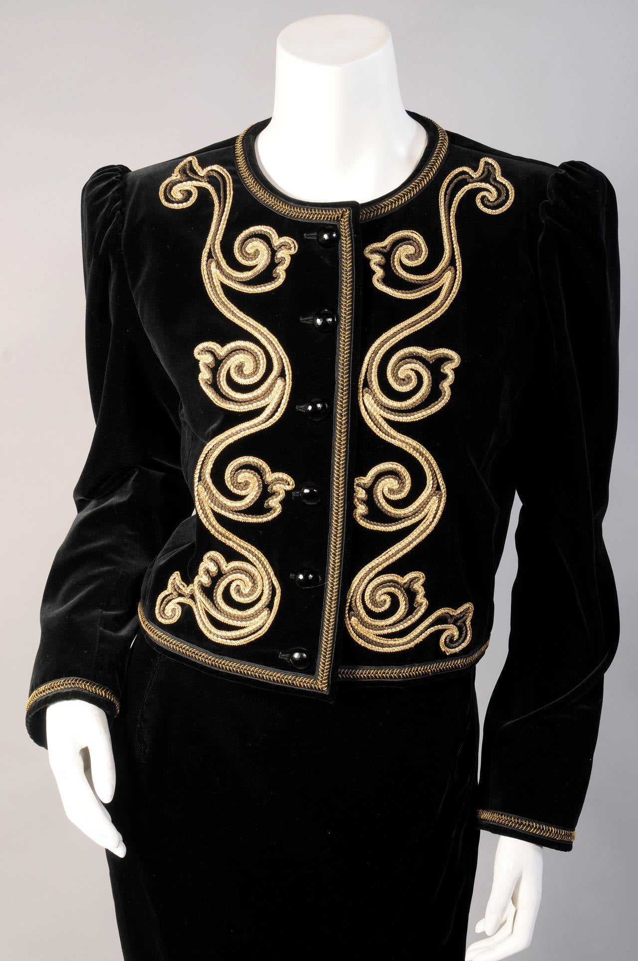 Rich gold soutache braid creates an elegant design on the front of this classic black velvet jacket. There is a second gold braid used to edge the jacket and cuffs, and it  closes with black jet buttons. The long lean matching skirt has a high slit