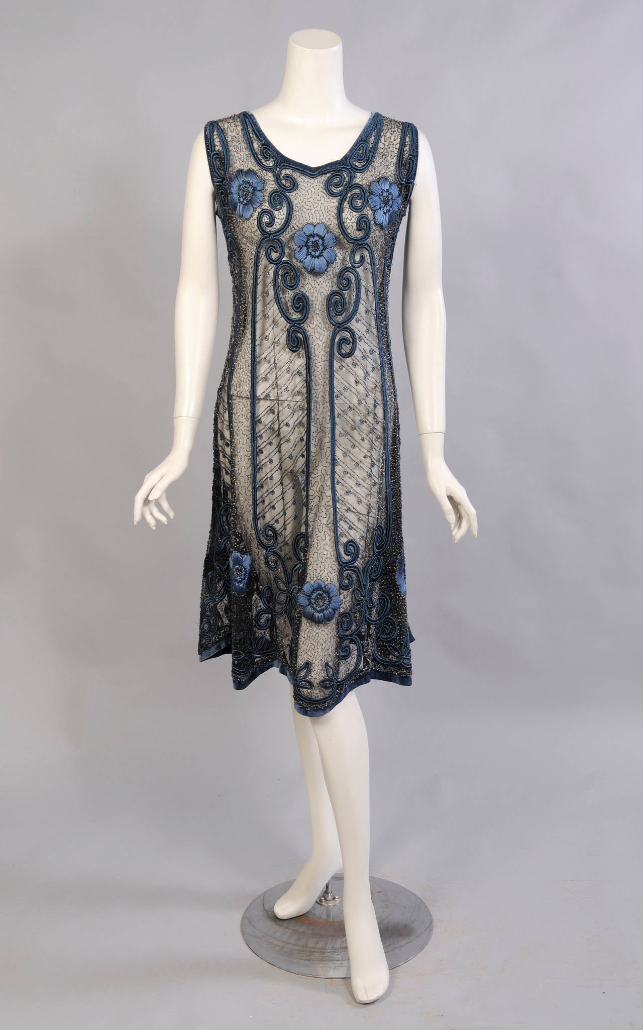 Fine black net is beaded with an all over vermicelli pattern using blue and clear caviar beads. The dress is embroidered with a matching blue silk floss for the flowers and scrolling designs. The neckline, arm hole and hem are bound in a matching