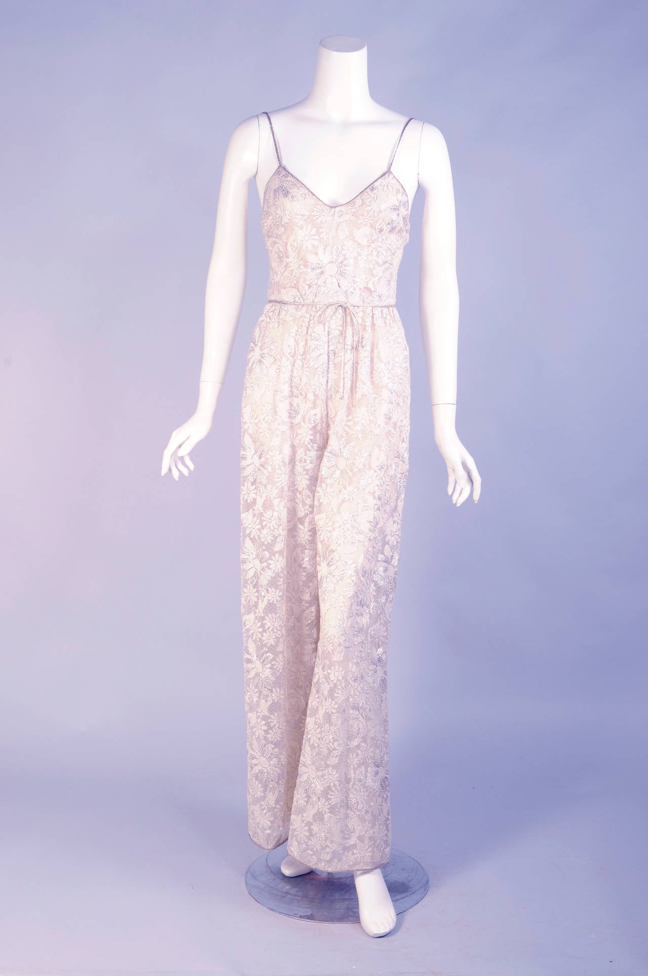 Sparkling silver floral lace is lined with a sheer nude silk chiffon for the jumpsuit of this ensemble. The neckline is edged with silver cord which is also used for the narrow straps, tie belt and the pant hem. The jumpsuit closes at the center
