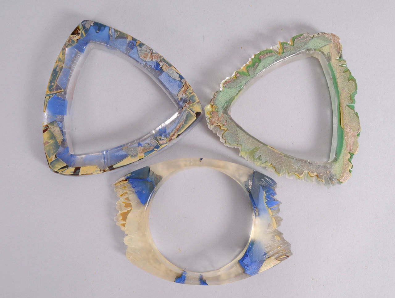 These unusual art glass bracelets are carved and polished to show off all of the gorgeous colors. Done in shades of blue and green they are reminiscent of beach glass. Two are shaped like a triangle and the oval one is signed. They are in excellent