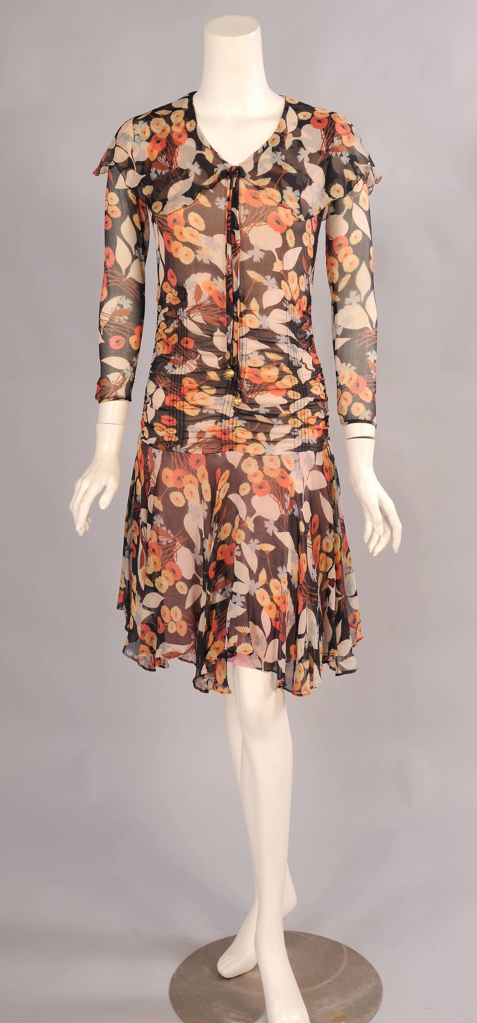 A black silk chiffon is printed with bouquets of orange and yellow flowers as well as a larger scale white and blue floral pattern. The dress has a sailor collar  and a bow with golden beads at the center front. The bodice is shirred over the hips