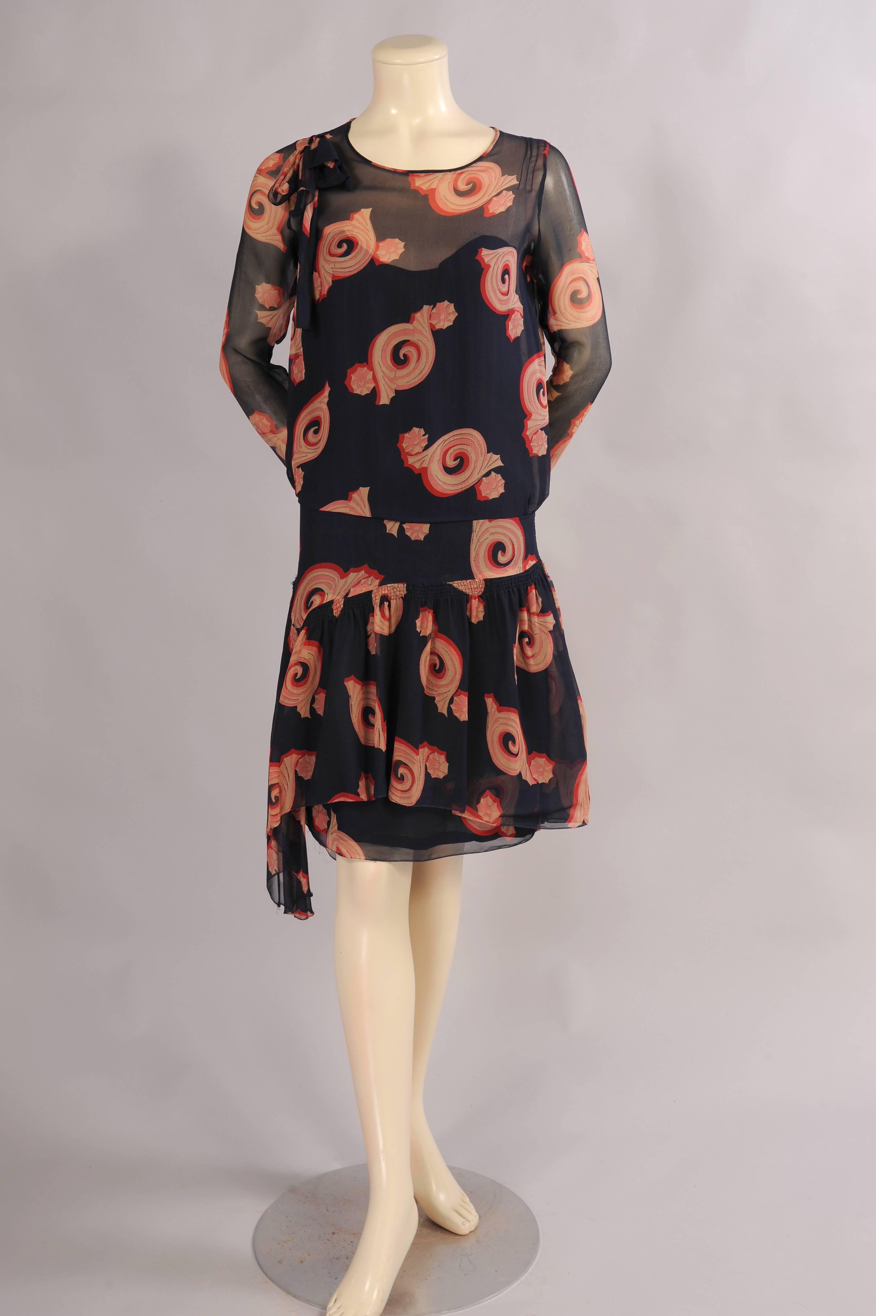 Navy blue silk chiffon is printed with an Art Deco design in red, pink and beige.
The dress has a bow on the right shoulder, long sleeves, a dropped waist  with a wide flat panel over the hips and a gathered skirt that dips lower on the right side.