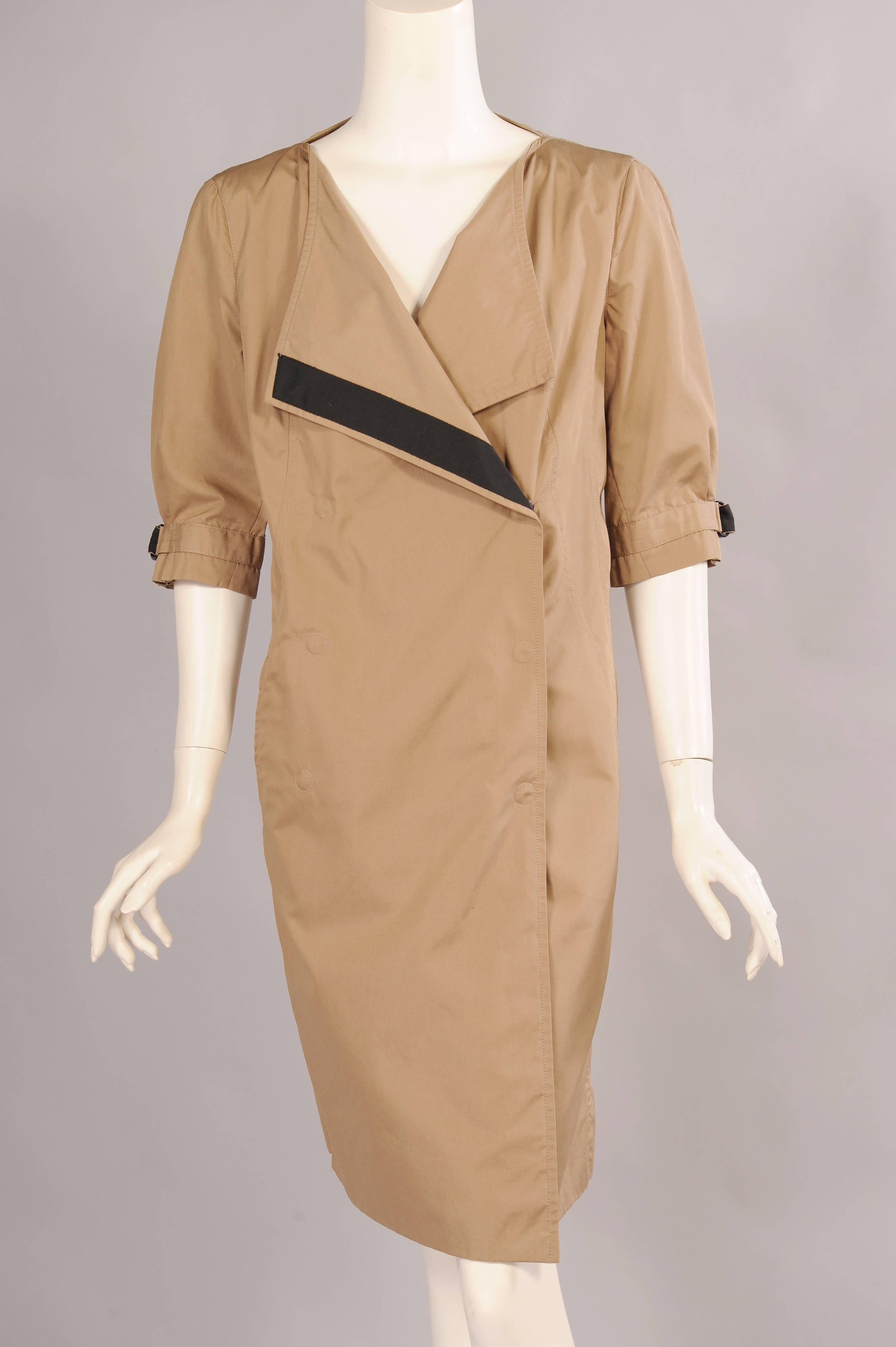 A classic khaki colored cotton day dress is updated with an open neckline, black grosgrain ribbon trim, and a concealed snap closure. The dres has elbow length sleeves with D Rings echoing the closure on the belt. Two pockets are concealed in the