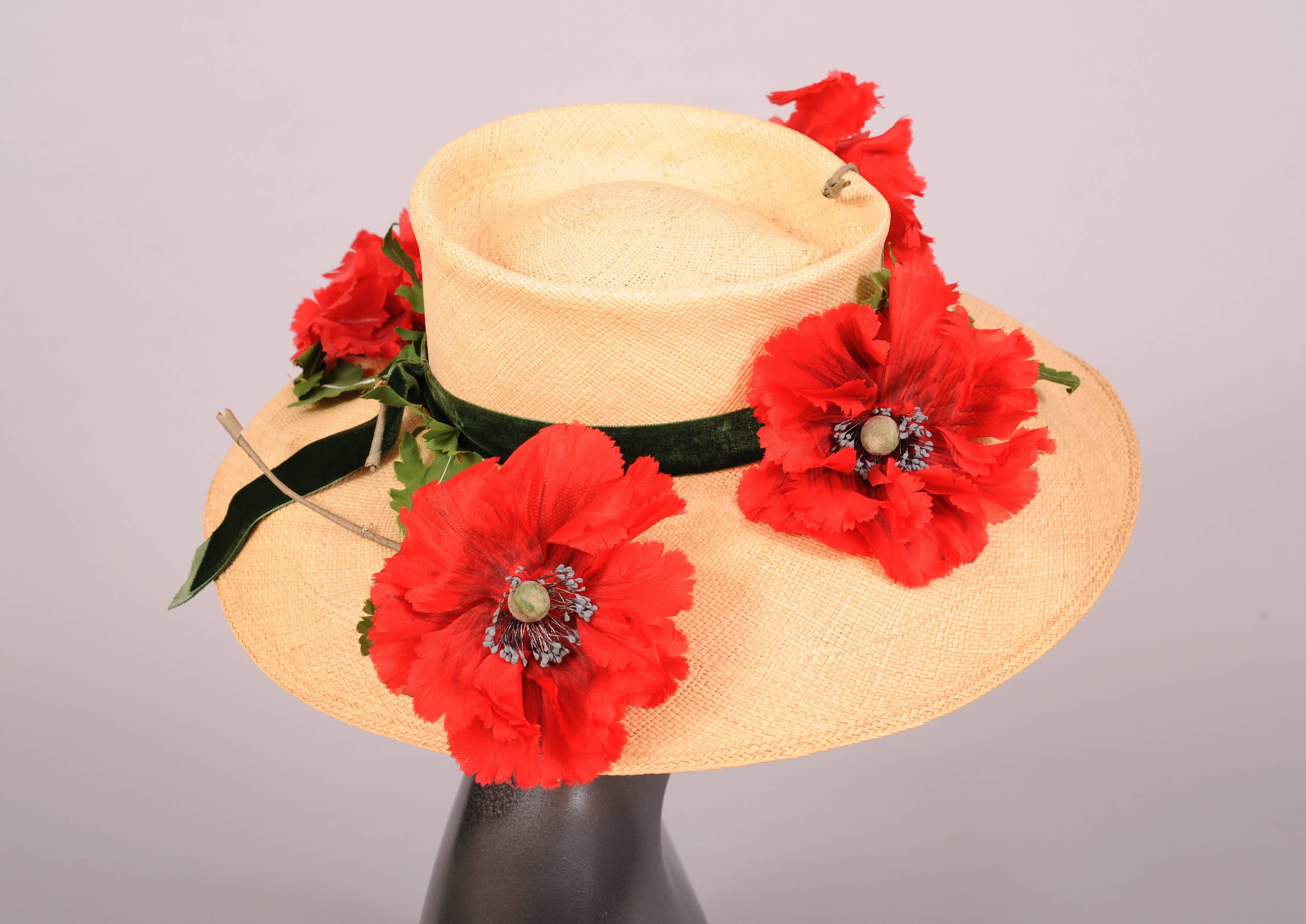 A finely woven natural straw hat with a wide brim is trimmed with a green velvet ribbon and luscious red silk poppies. They are scattered around the brim in a random pattern and they are accented with green leaves. The hat is in excellent