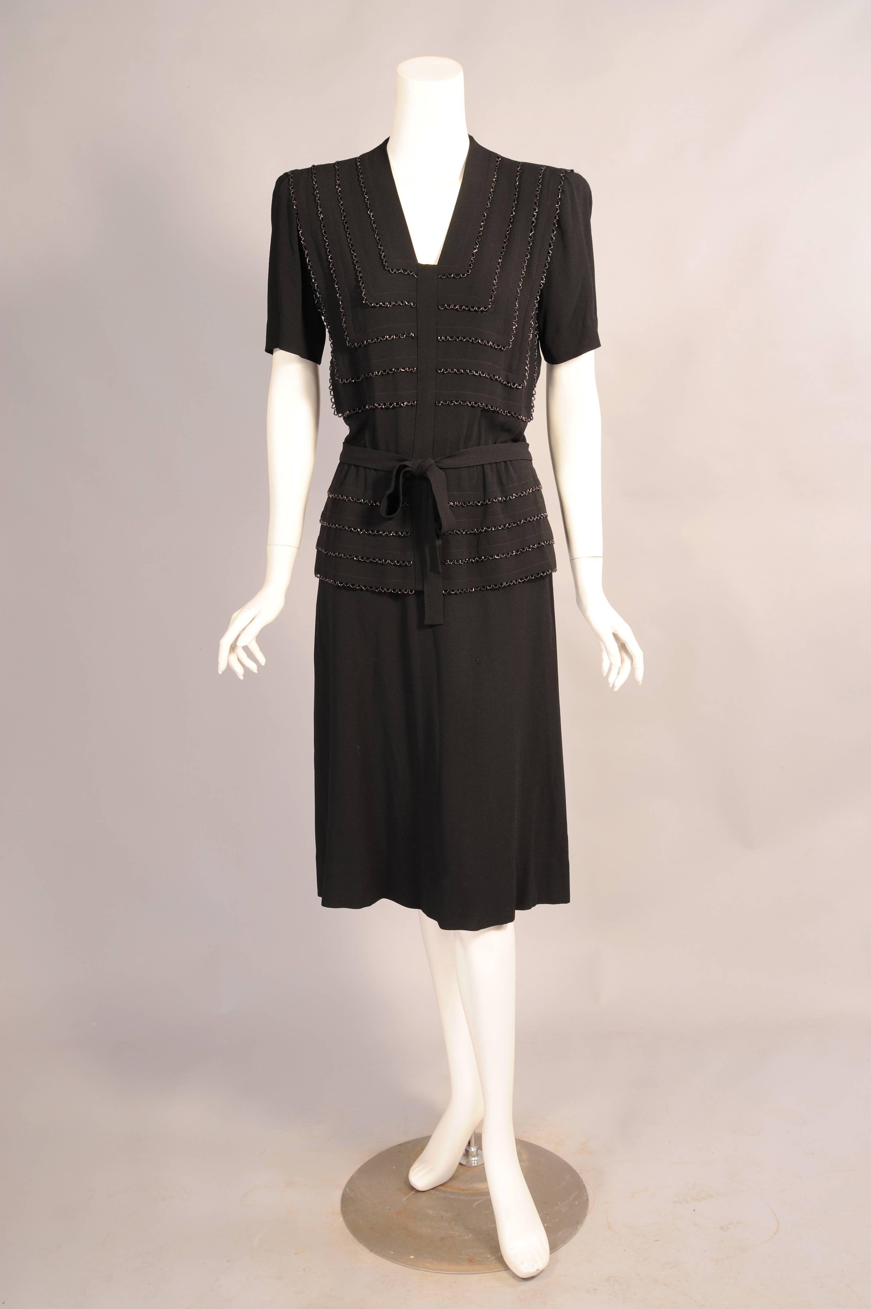 This elegant black crepe dress from the 1940's has a V shaped neckline, short sleeves and a natural waist with self tie. The bodice and peplum are horizontally pleated and these pleats are edged with scalloped black caviar bead work. The dress has
