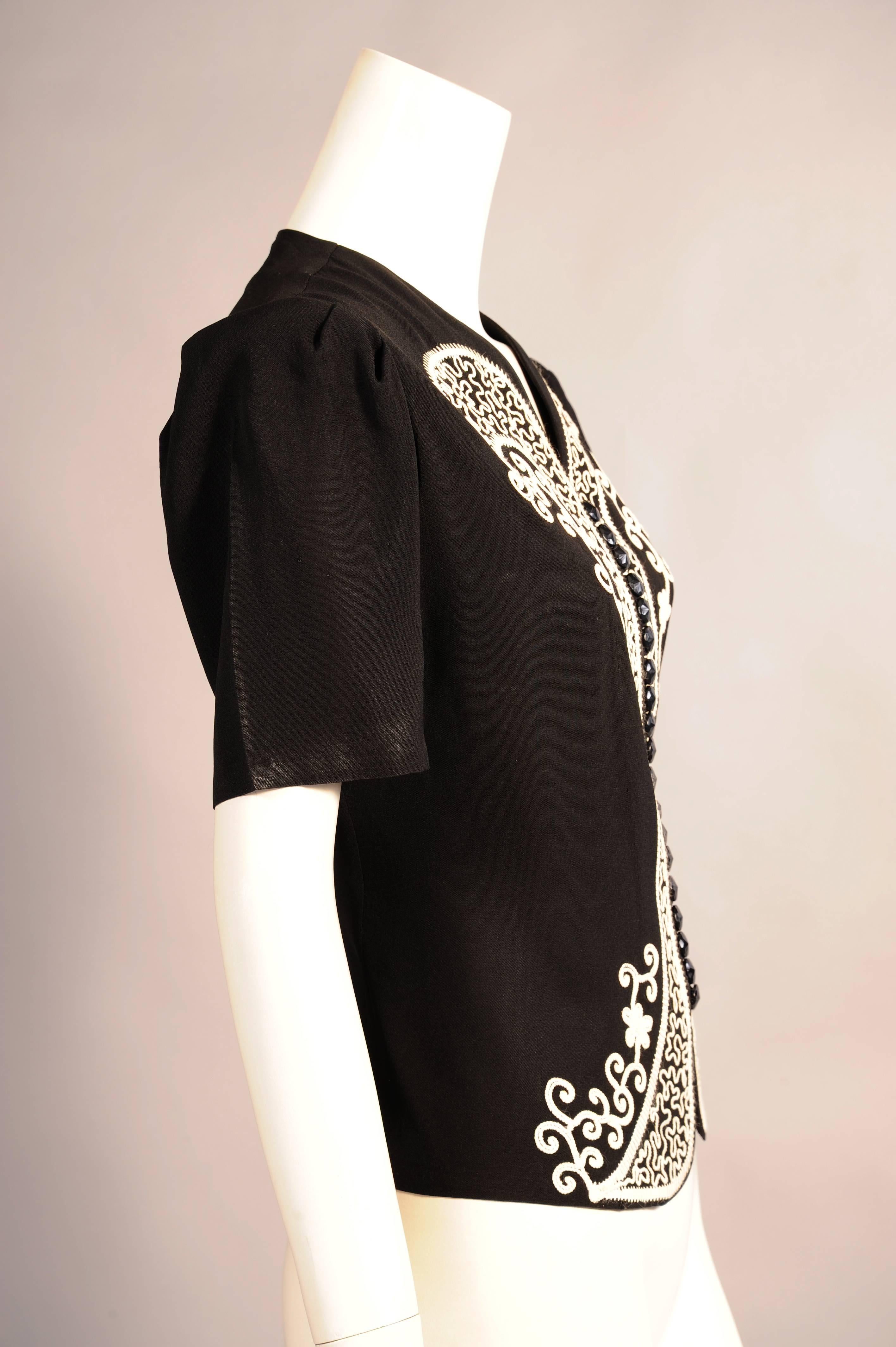 This short sleeved jacket black has lavish white soutache braid trim on both sides of the center front. In between there is a row of twenty black jet buttons with fabric loops.  This graphic jacket is in excellent
