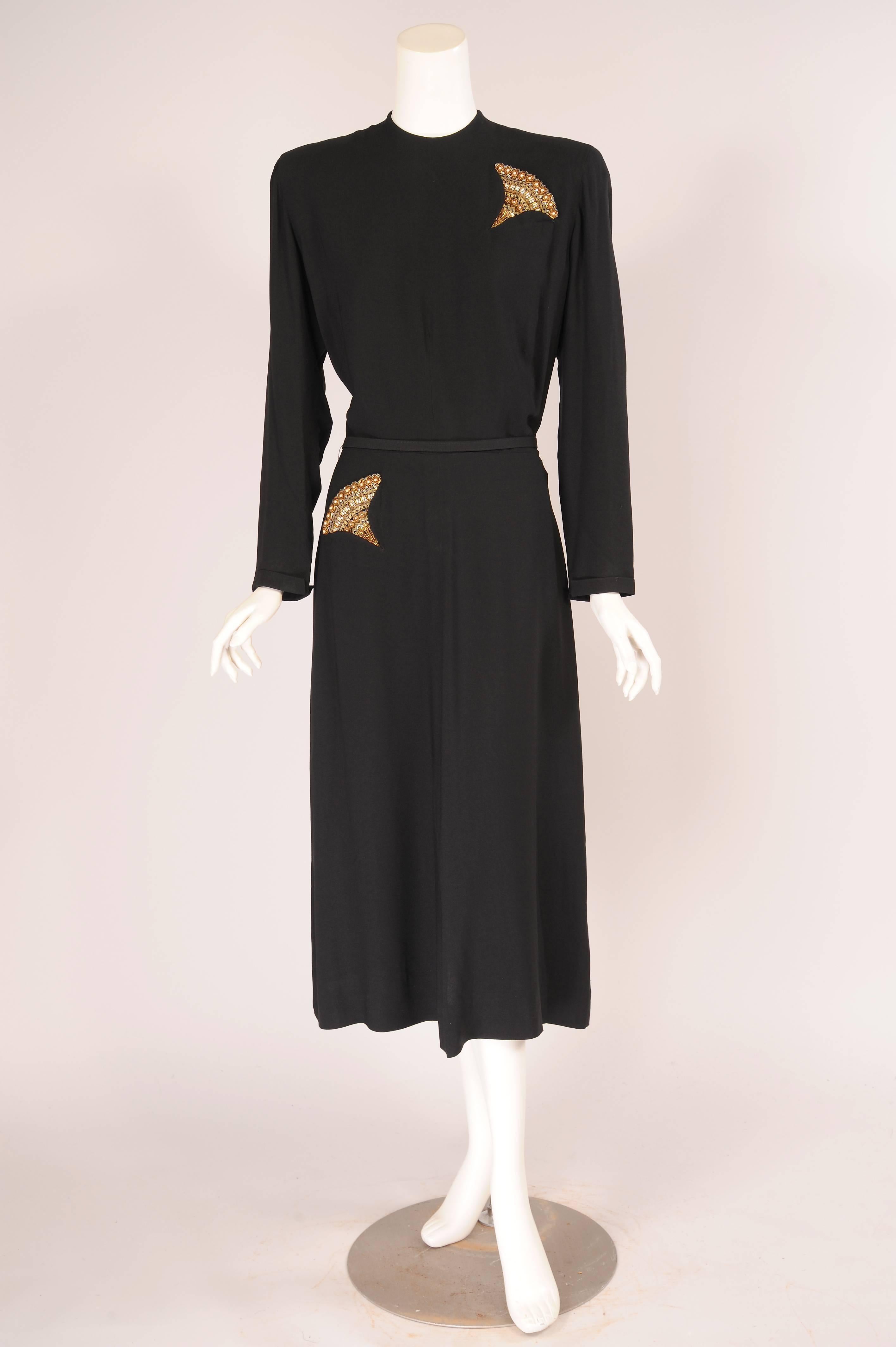 Gold braid, pearls and rhinestones are used for the decorative fan shaped elements emerging from the pockets of this larger size black crepe dress designed by Maurice Rentner. The dress has a dramatic drape at the center back of the mid calf length