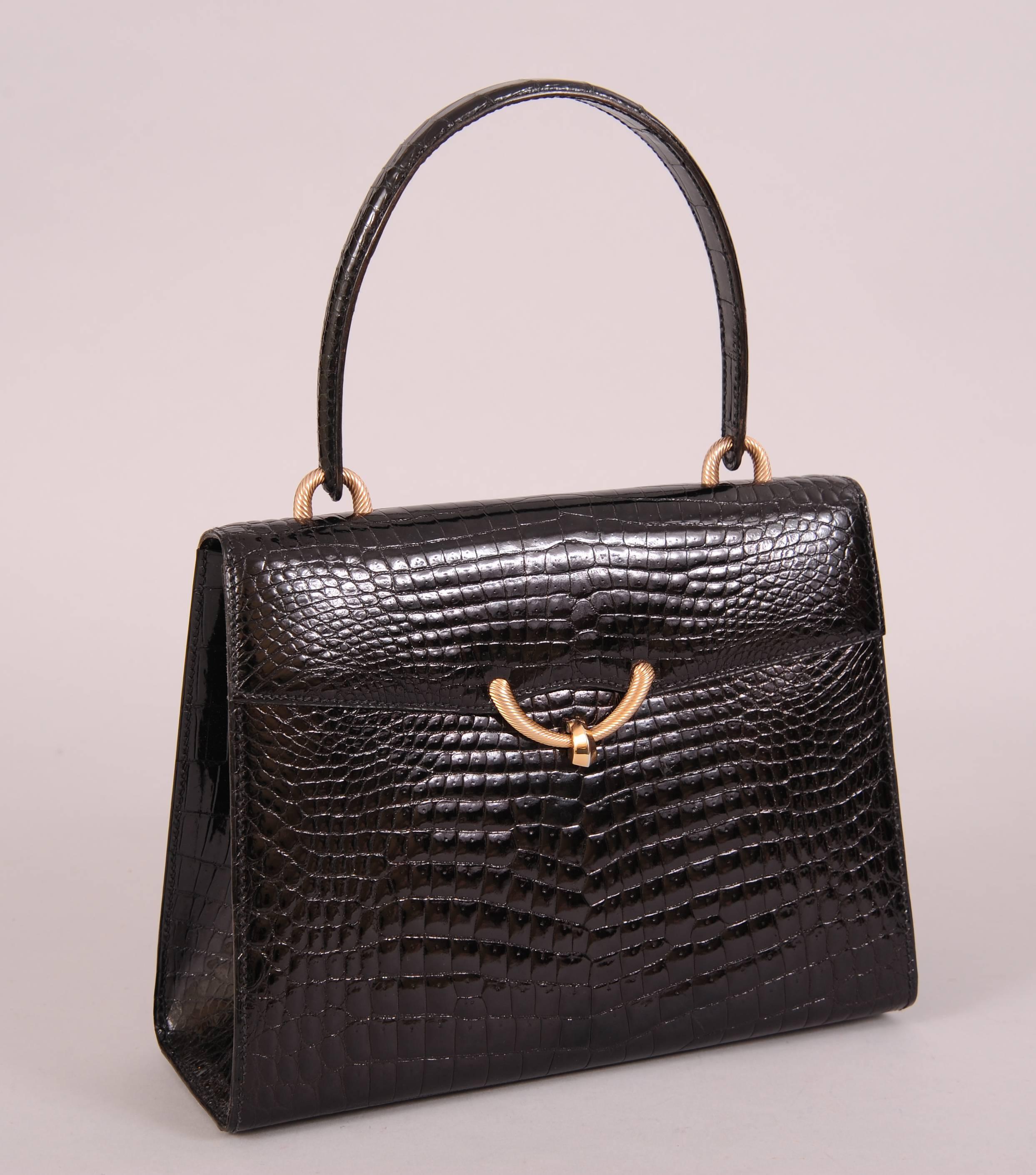A structured black crocodile handbag with a lustrous shine is accented with gold toned hardware. There is a large slip pocket on the front of the bag concealed by the flap closure. There are two more slip pockets, a zippered pocket and a metal