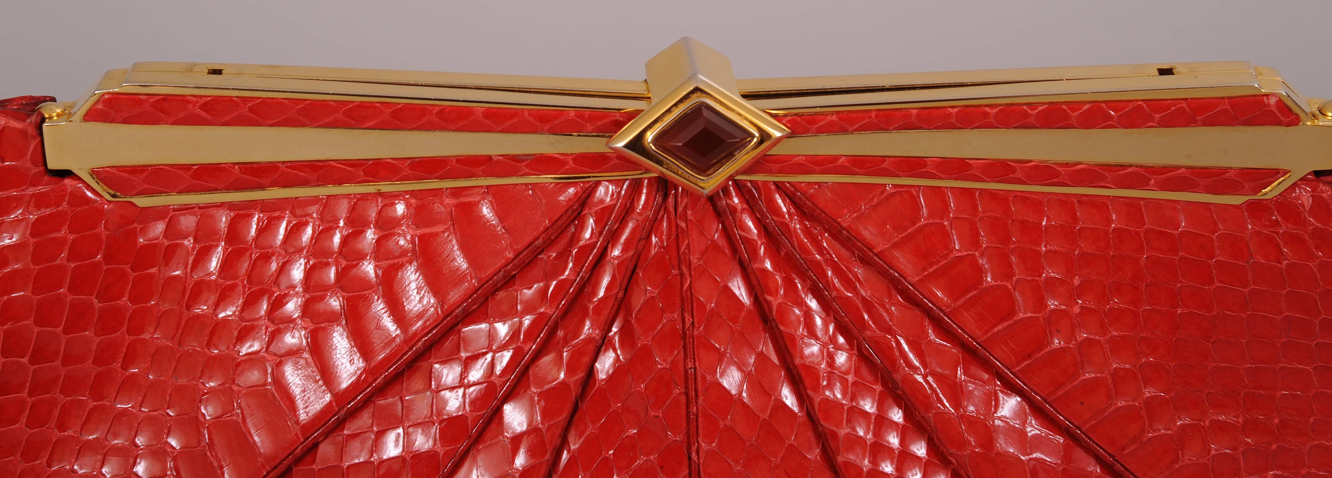 A tailored bag made form Bright red snakeskin is pleated on the front and back. The hinged frame is gold toned metal and red snakeskin with a faceted stone clasp. The bag has a gold toned chain strap tucked inside. The red satin lining is in perfect