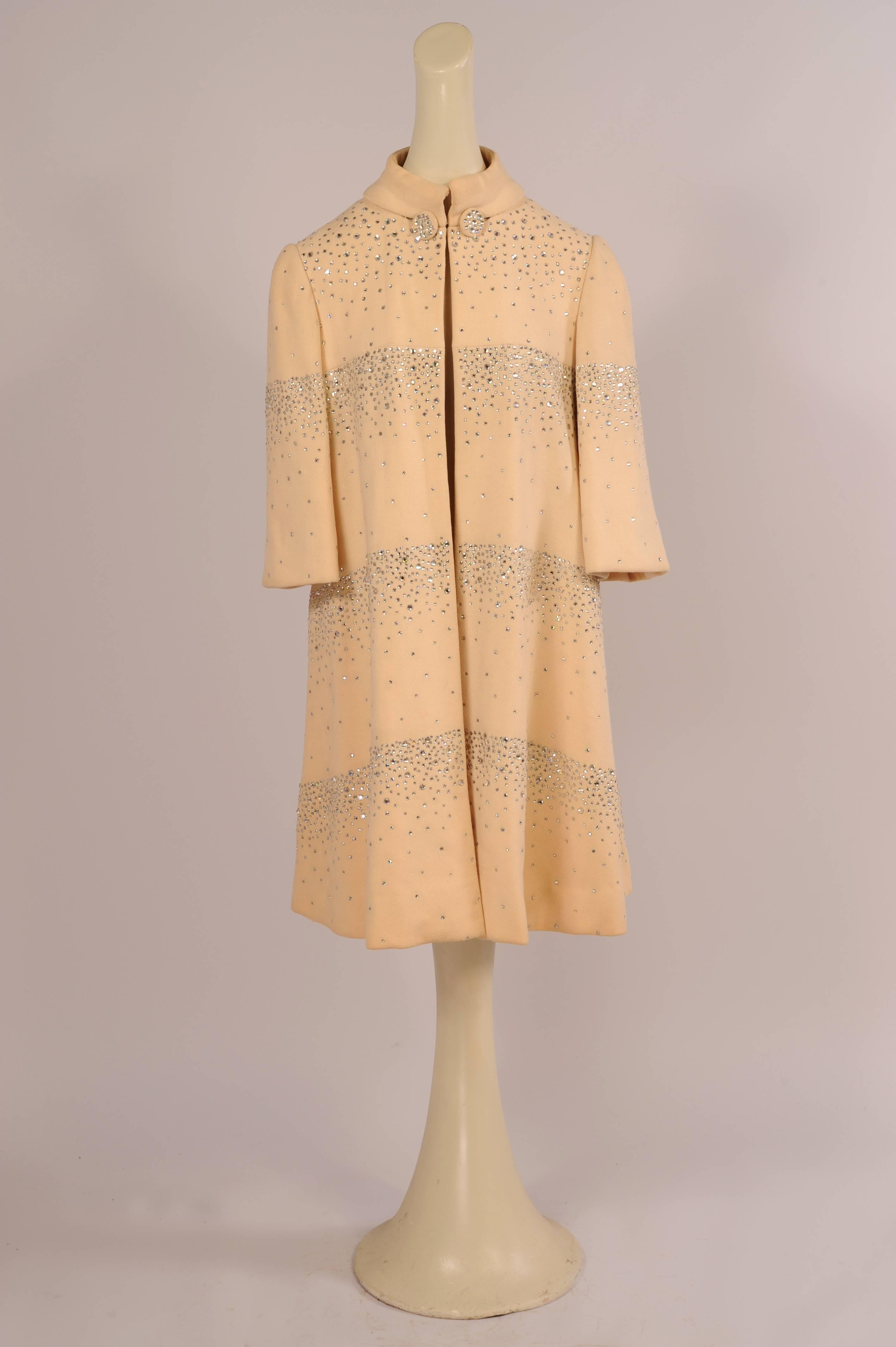 Sparkling and eye catching, this is a rare cream colored wool crepe example of this stunning coat designed by Pauline Trigere. It is covered with prong set rhinestones in various sizes. They begin as horizontal bands on the front of the coat and