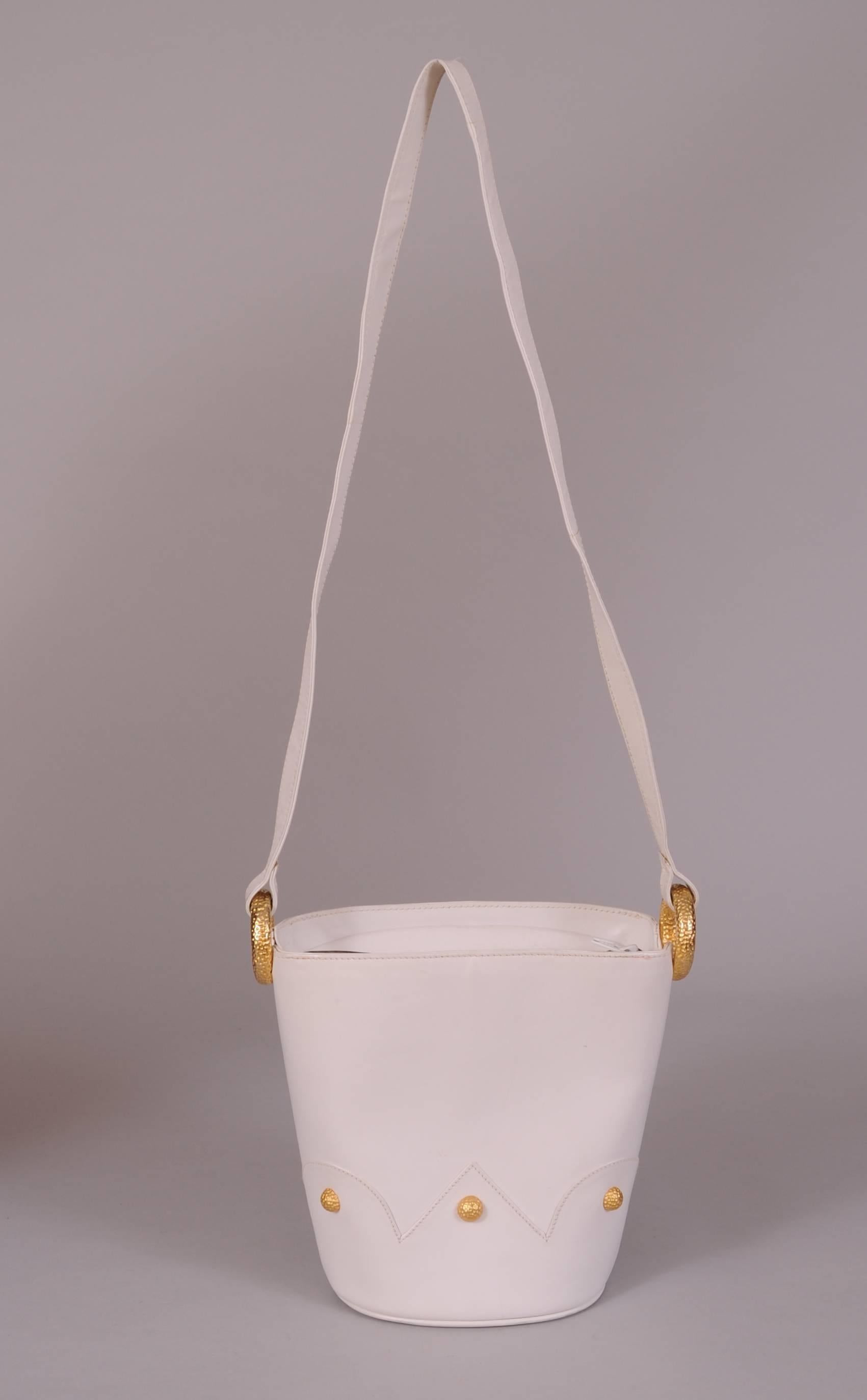 This charming white leather bucket bag from Paloma Picasso has a crown shaped applique all around the bag. It is studded with hammered gold toned
metal domes. These match the rings used to hold the shoulder strap. The bag has a zippered top opening,