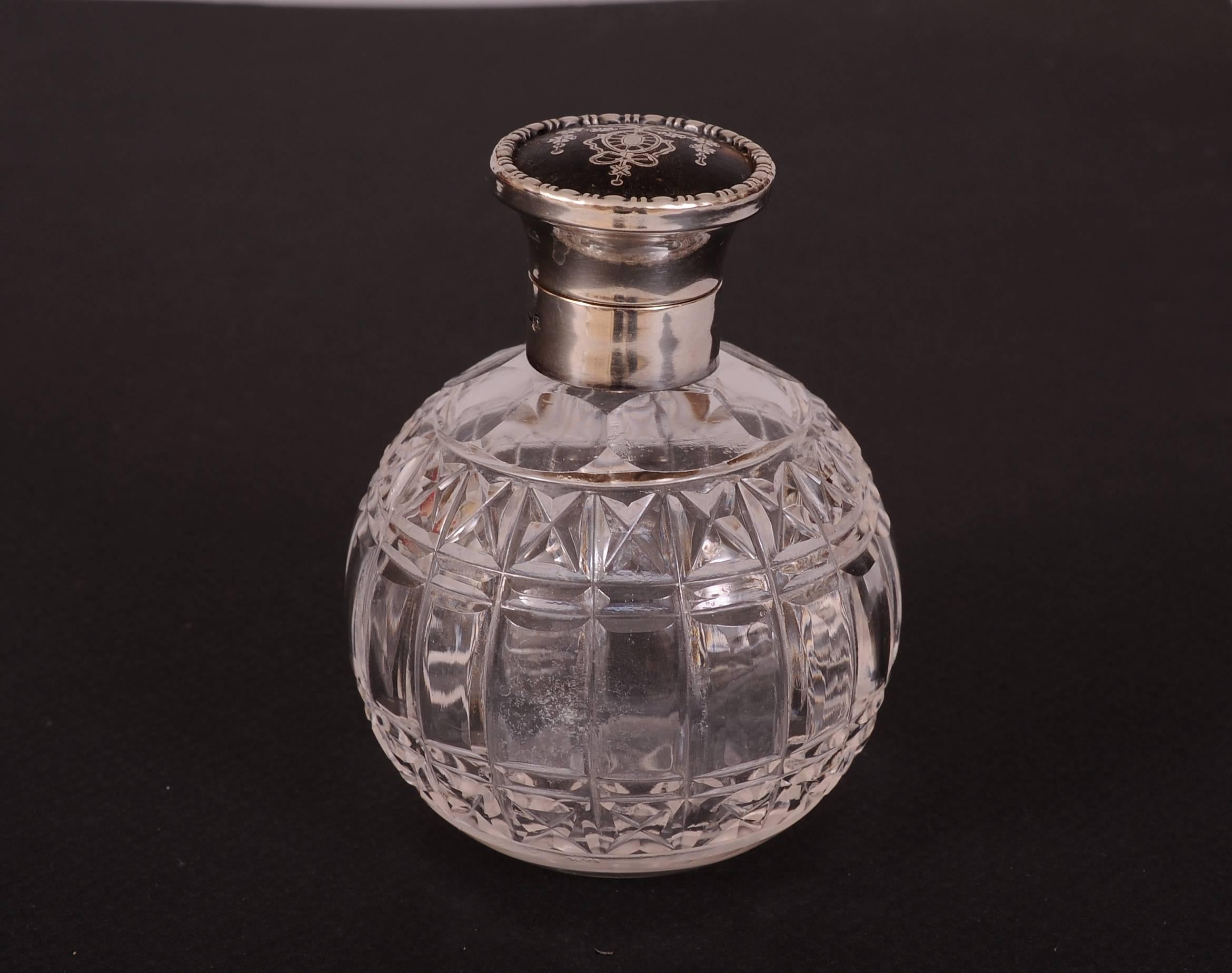 A hinged sterling silver top has a piece of tortoise shell inlaid with a sterling silver design of ribbons and flowers above a round cut glass bottle. The top is marked L&S followed by hallmarks. It is in excellent