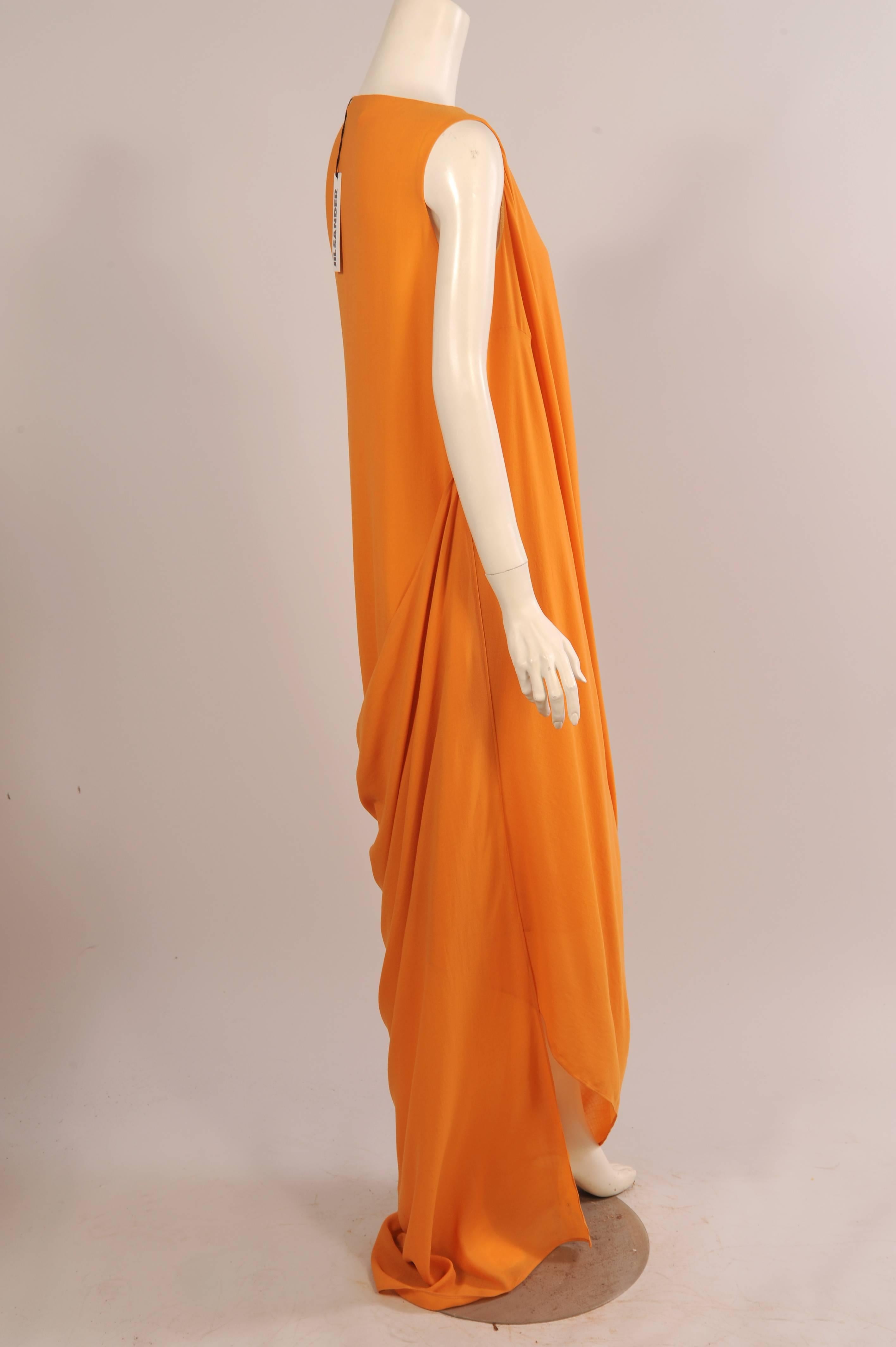 A classic knee length sleeveless sheath dress with a hidden left side zipper is transformed by a draped panel attached at the right shoulder seam. This follows the curve of the armhole and is attached all the way down to the hem of the dress on the