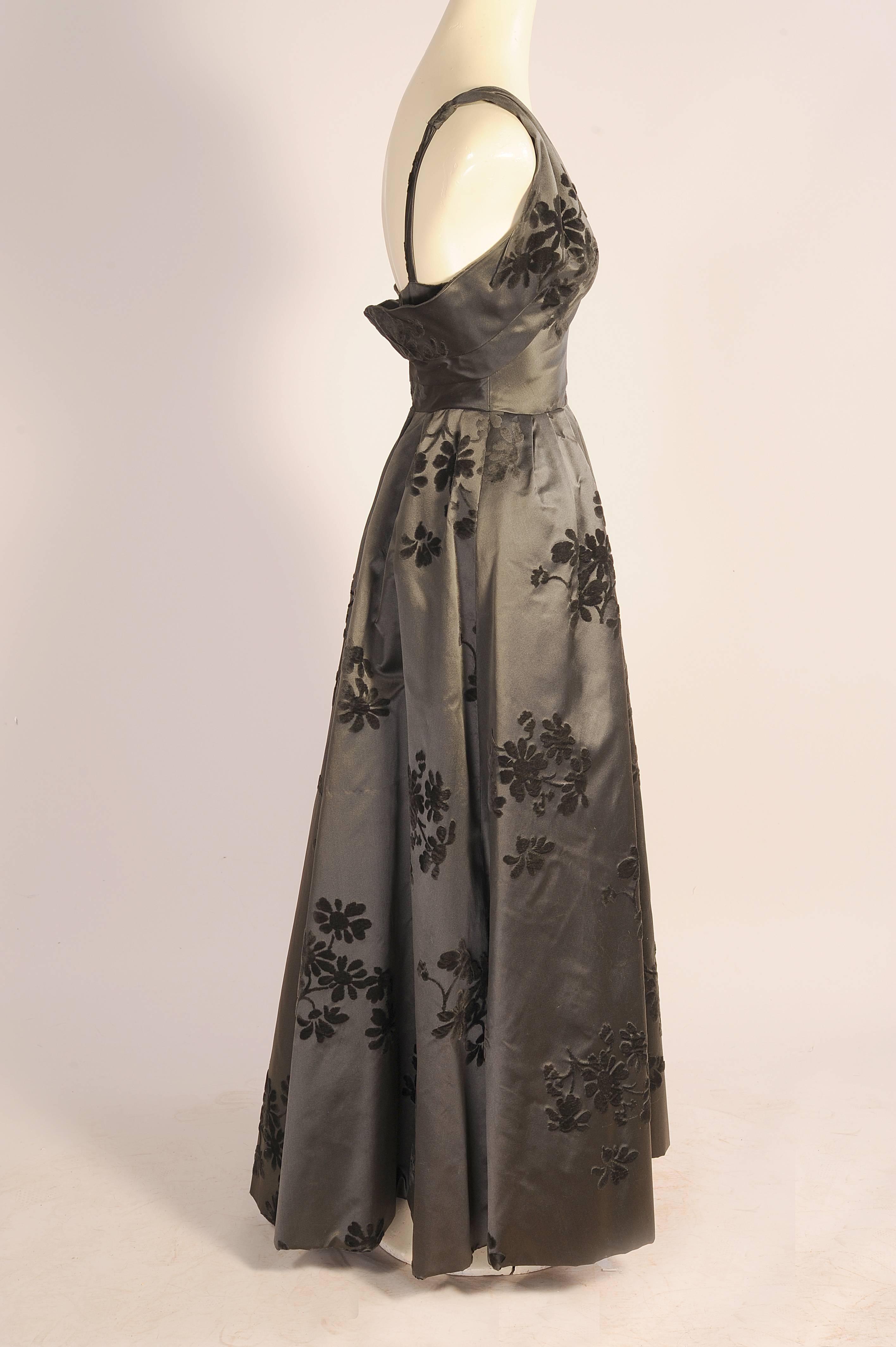This elegant evening gown is the perfect jewelry dress, so classic it is just waiting for your most glamorous gems.
It is made from charcoal grey silk satin with black velvet floral motifs throughout. The bodice is fitted with a V neckline and