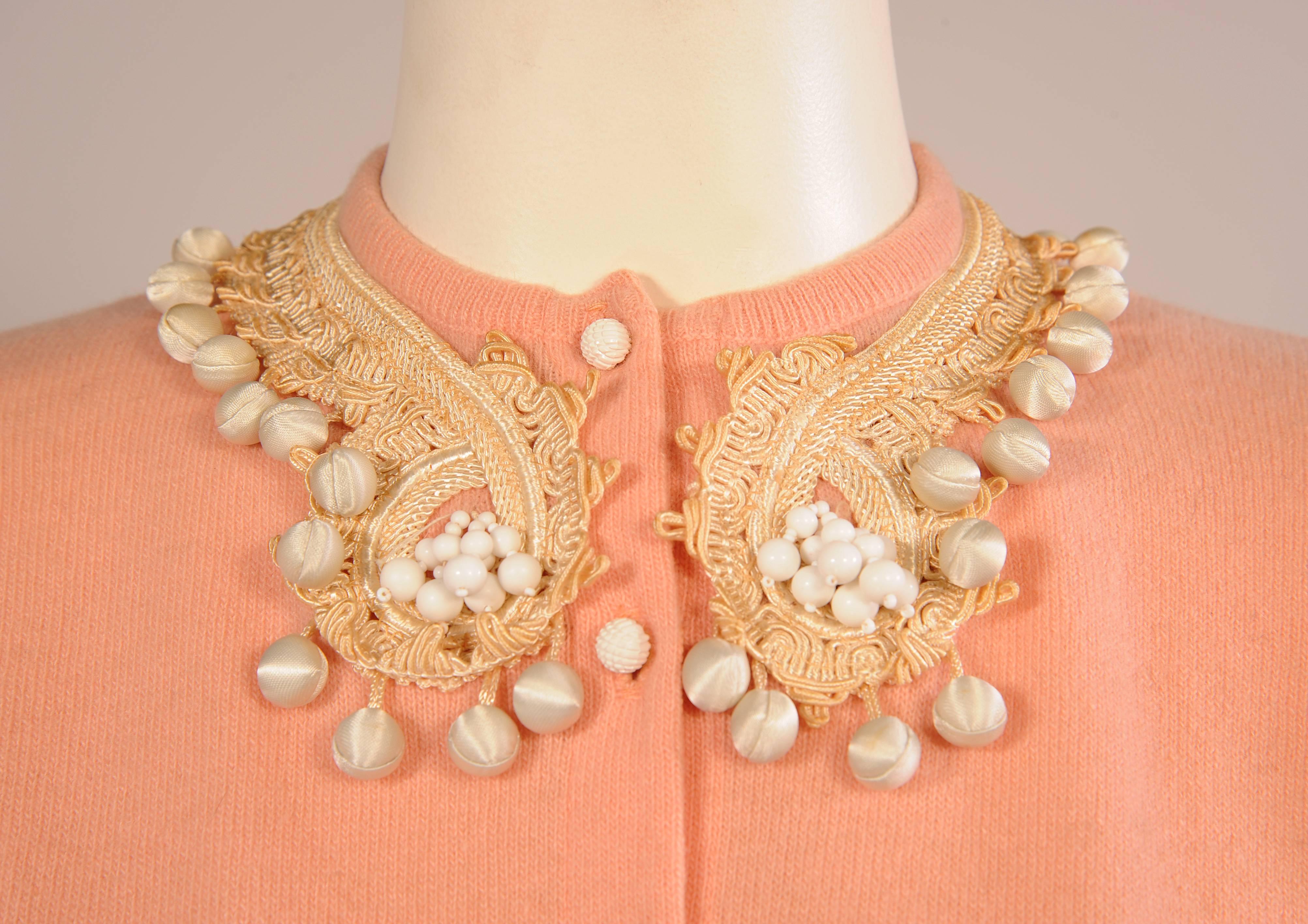 This sweet 1950's cashmere sweater is embellished with braid trim at the neckline and cuffs. A large cluster of white beads, en tremblant, is set into the turn of the collar trim. This is further enhanced with dangling white satin covered balls on