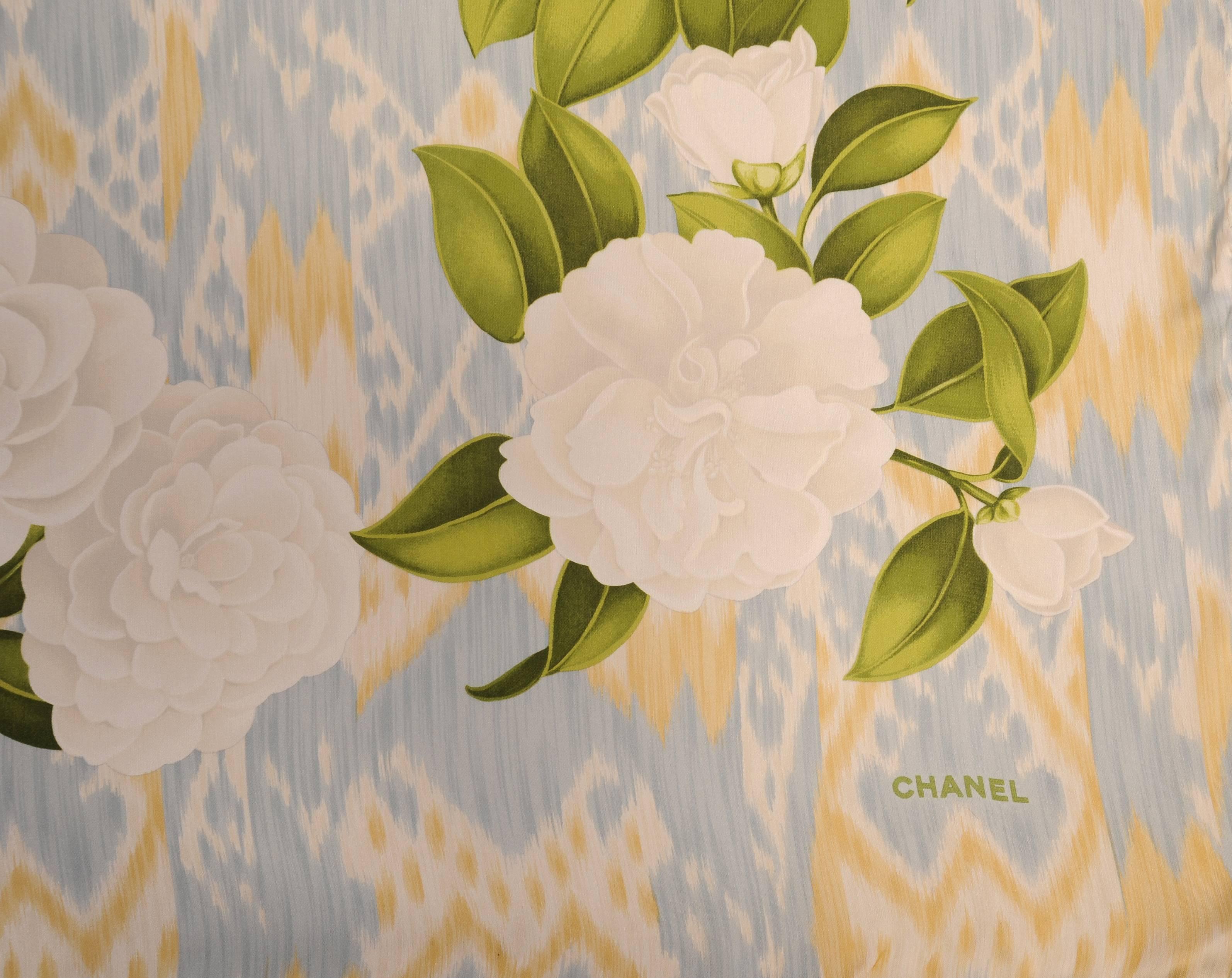 A summer weight silk scarf is strewn with white camellias and green leaves on a light blue, yellow and white Ikat pattern background. It is marked Chanel in one corner and still retains the original Saks Fifth Avenue tag and box. It is in excellent