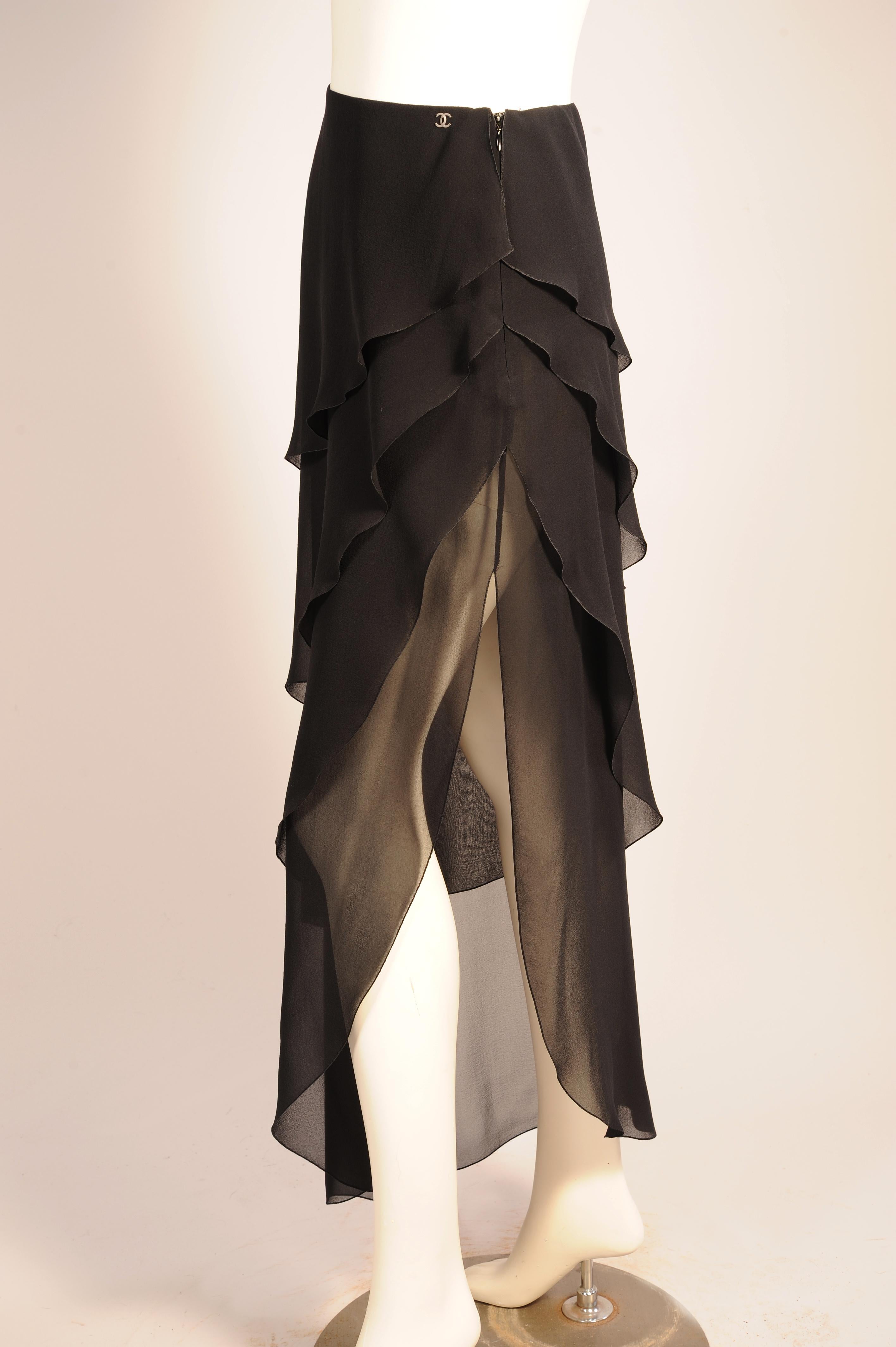 Quattro strati a forma di petalo di chiffon di seta nero scendono a cascata sui fianchi e cadono con grazia fino all'orlo curvo. L'apertura del lato sinistro  su questa gonna lunga di Chanel lascia intravedere le gambe. La gonna ha una fascia