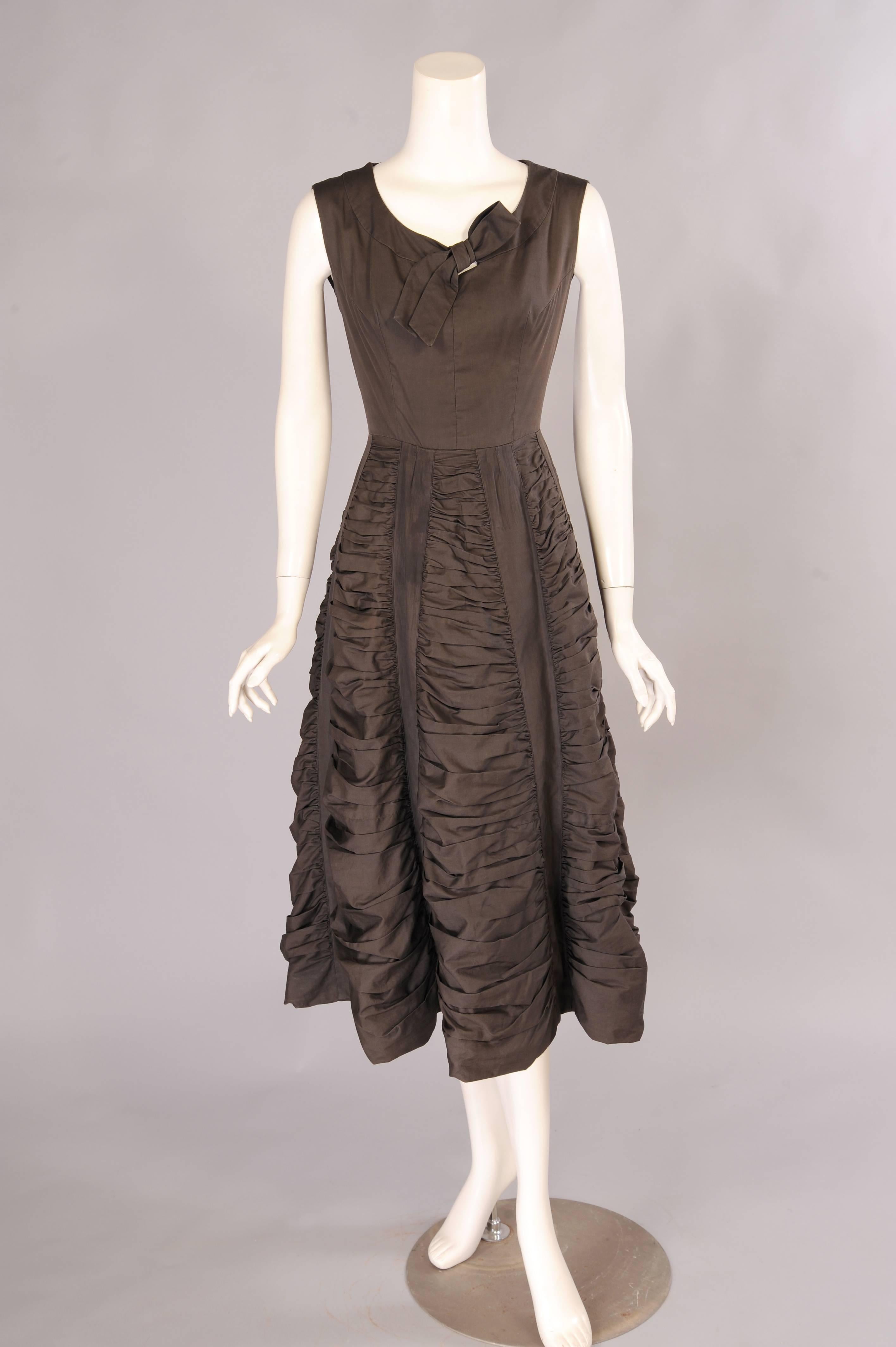 Dark chocolate brown cotton sets this 1950's cocktail dress apart from the LBD. It has a faux tie neckline, fitted bodice and a full skirt. The skirt is gathered horizontally for added interest and fullness. The gathers are held in place by flat