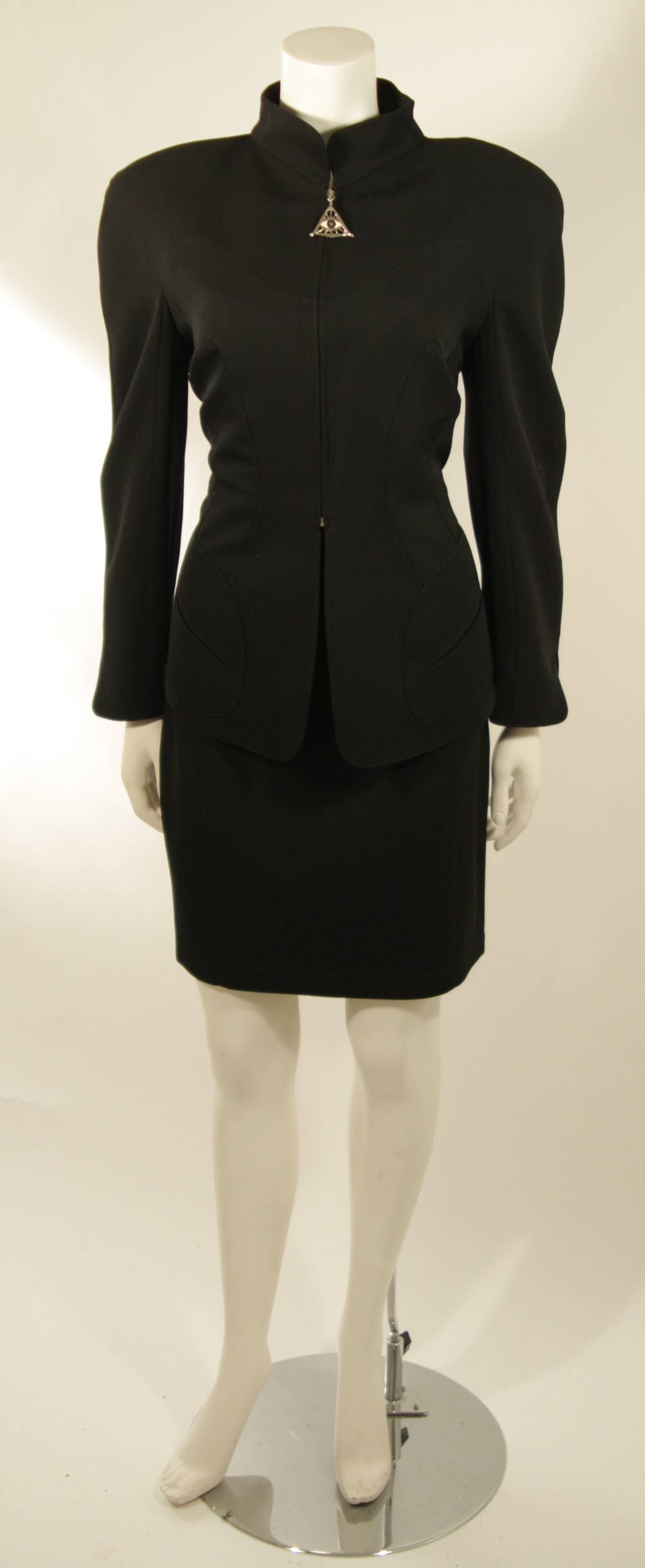 This Thierry Mugler design is available for viewing at our Beverly Hills Boutique. This two piece skirt suit is composed of a black fabric, and features a mandarin style collar which is accented by a 